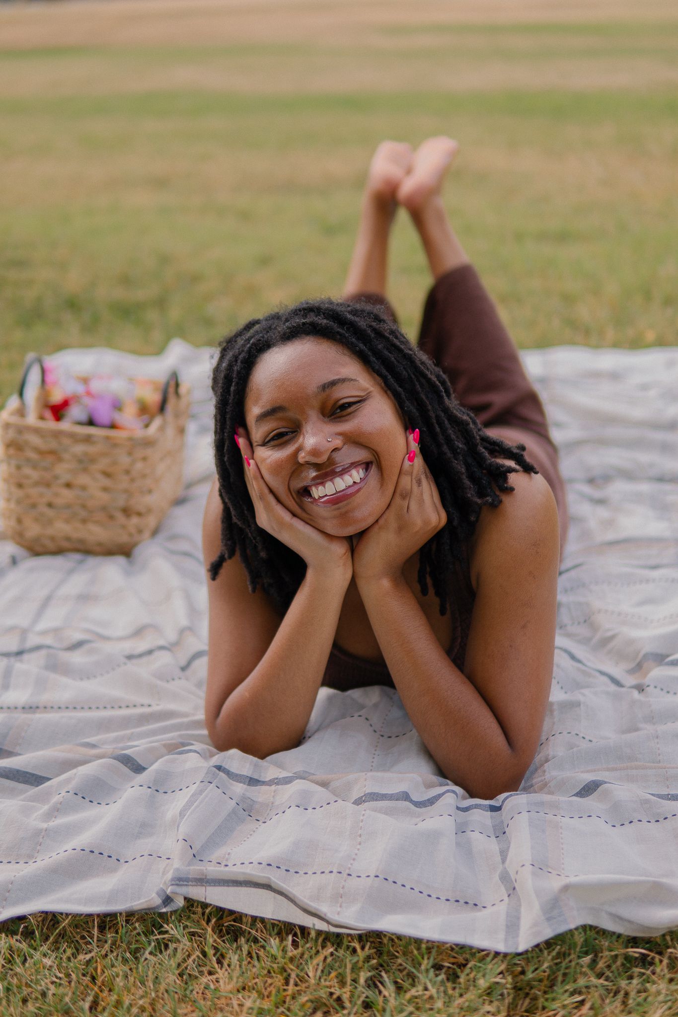 A person lying on a picnic blanket outdoors enjoying a sunny day with a basket of treats nearby.