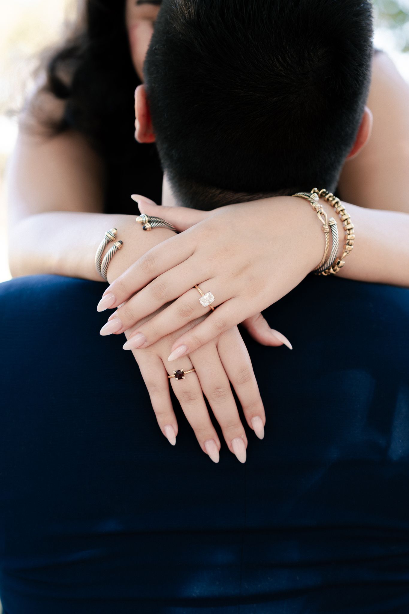 Close-up shot of hands with elegant manicure and engagement ring embracing against navy blue background.