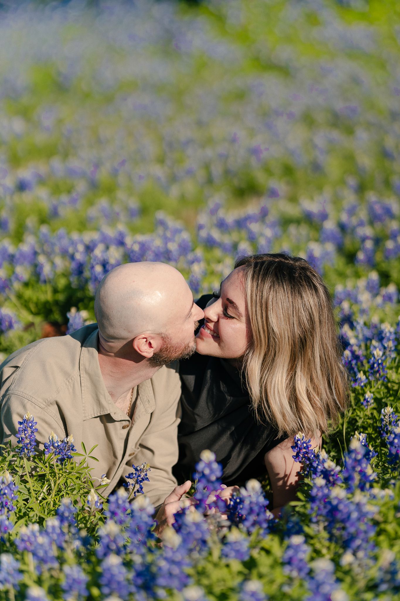 A couple shares a tender kiss while laying in a field of vibrant purple bluebonnets on a sunny spring day.