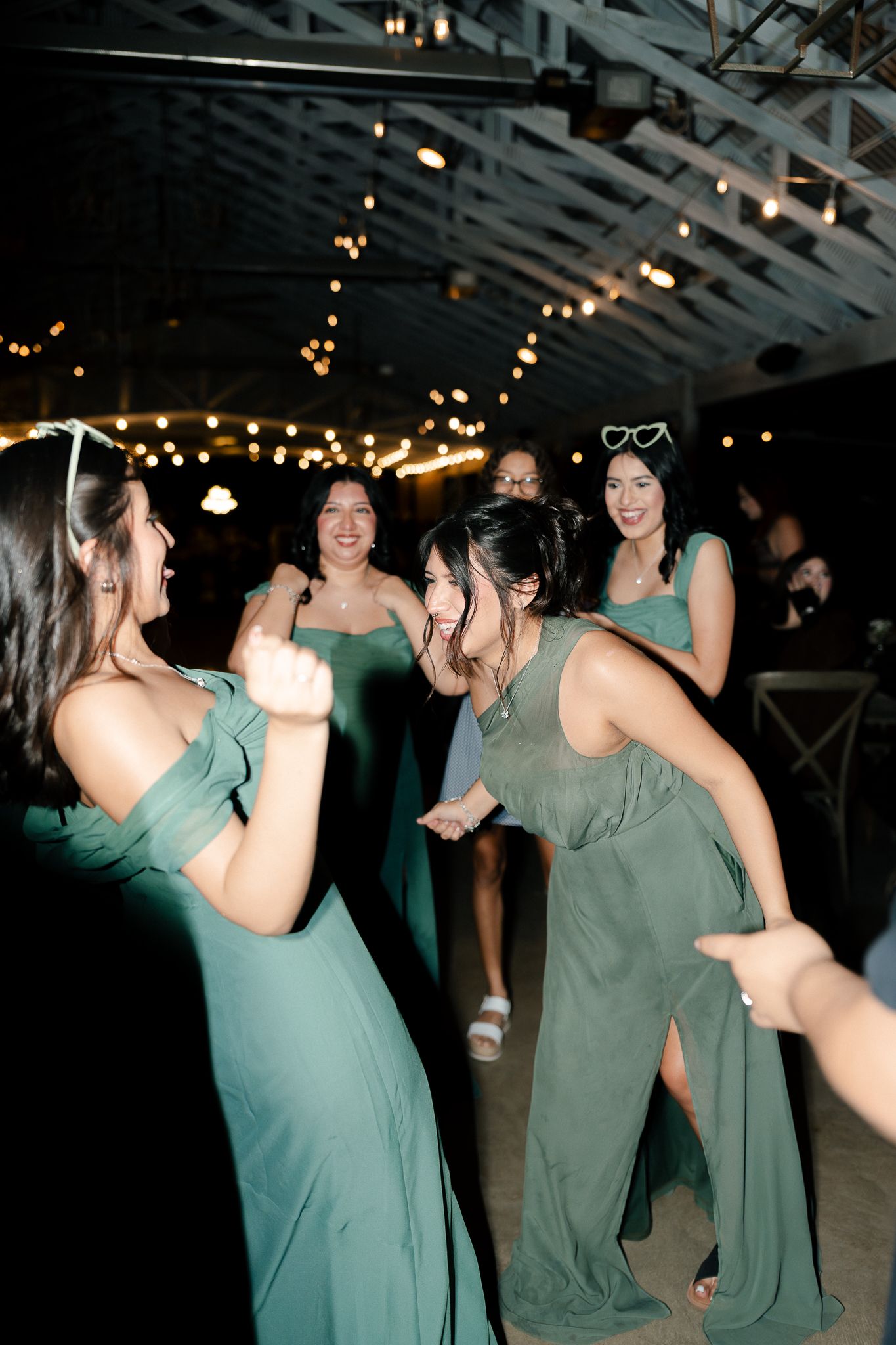 Bridesmaids in matching seafoam green dresses dance together under string lights at an evening wedding reception.
