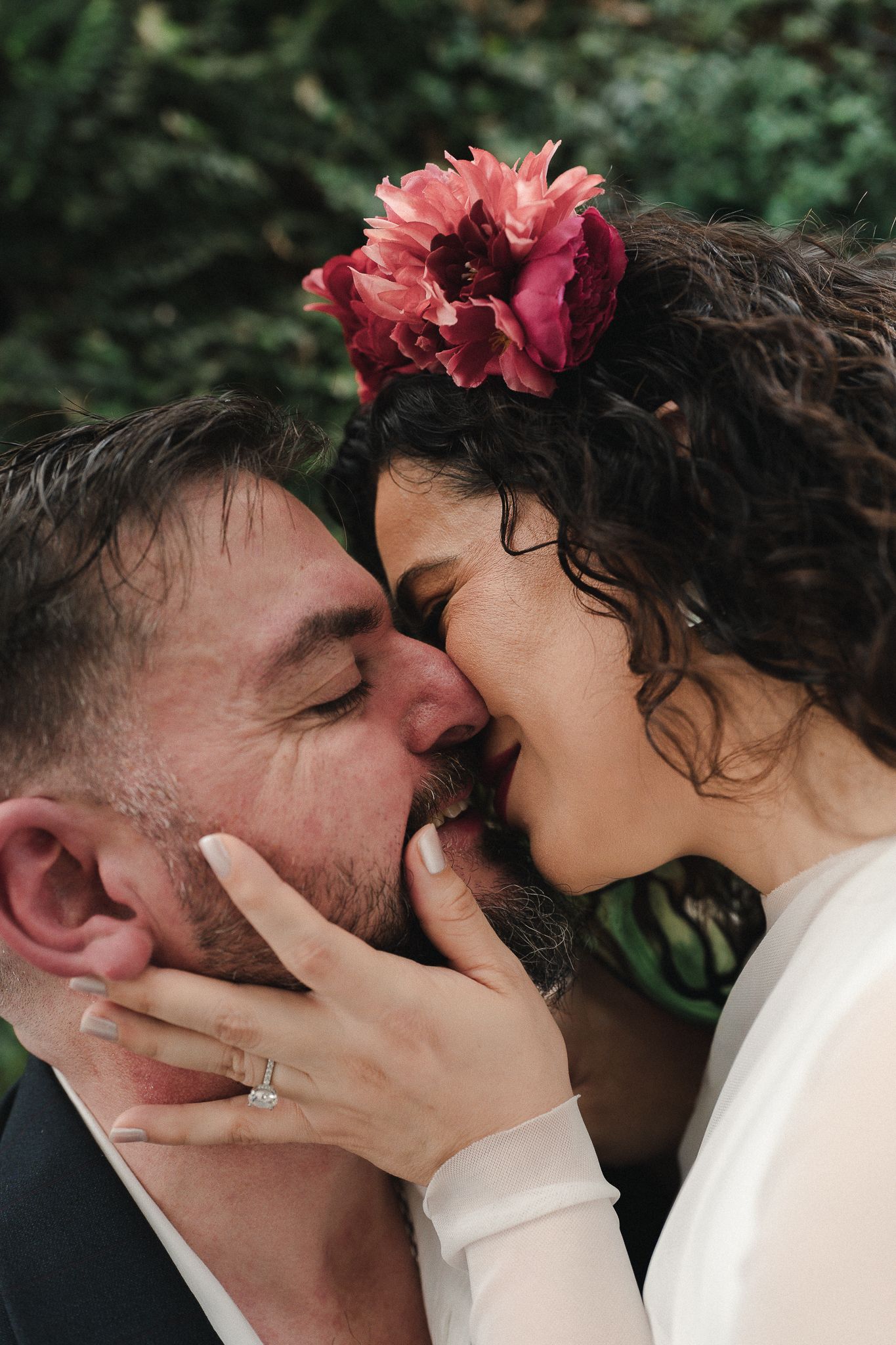 A couple shares a romantic kiss, bride wearing a red floral hair accessory and engagement ring, outdoors.
