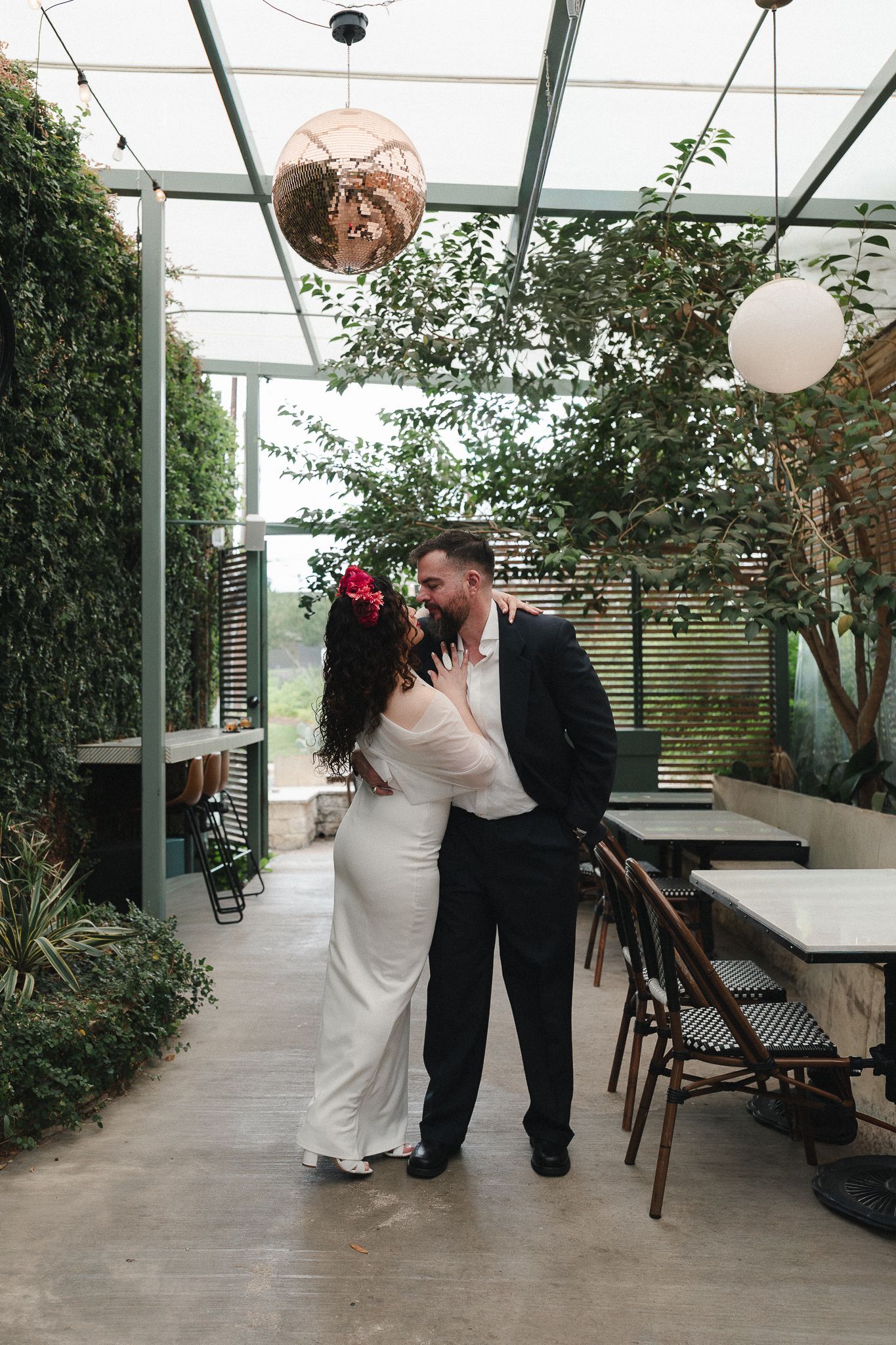 Bride and groom share a romantic moment in a greenhouse venue with lush greenery and stylish pendant lights.