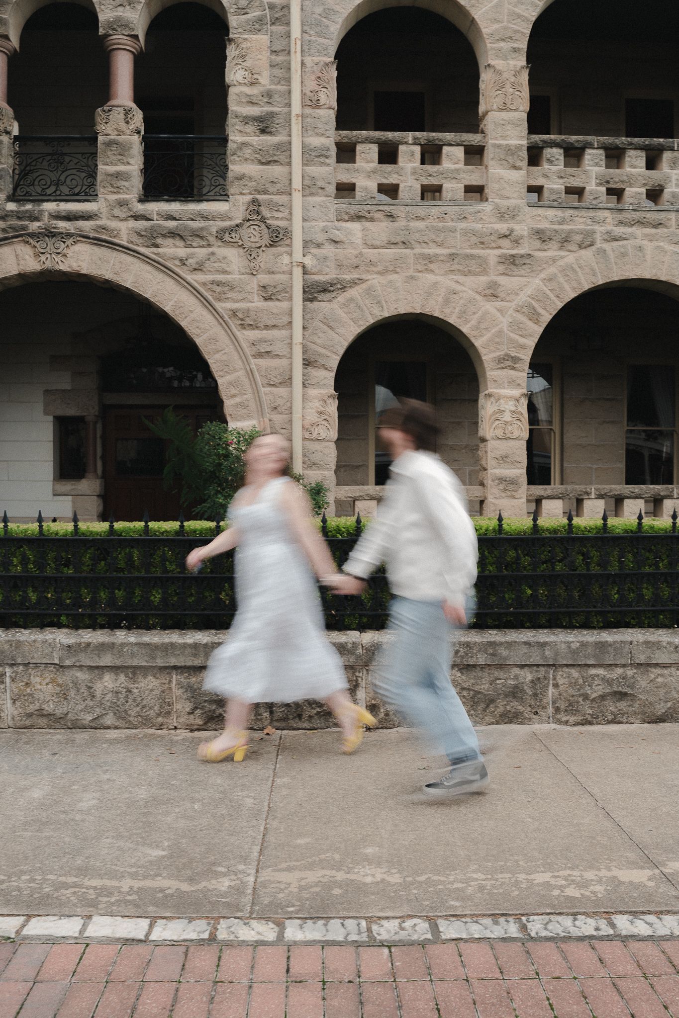 Blurred couple holding hands walking past historic stone building with arched colonnade.