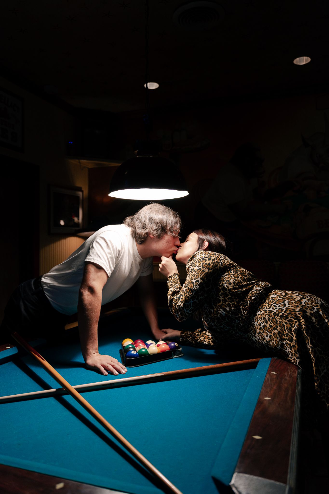 Couple sharing a romantic kiss over a pool table with billiard balls and cues under dim light.