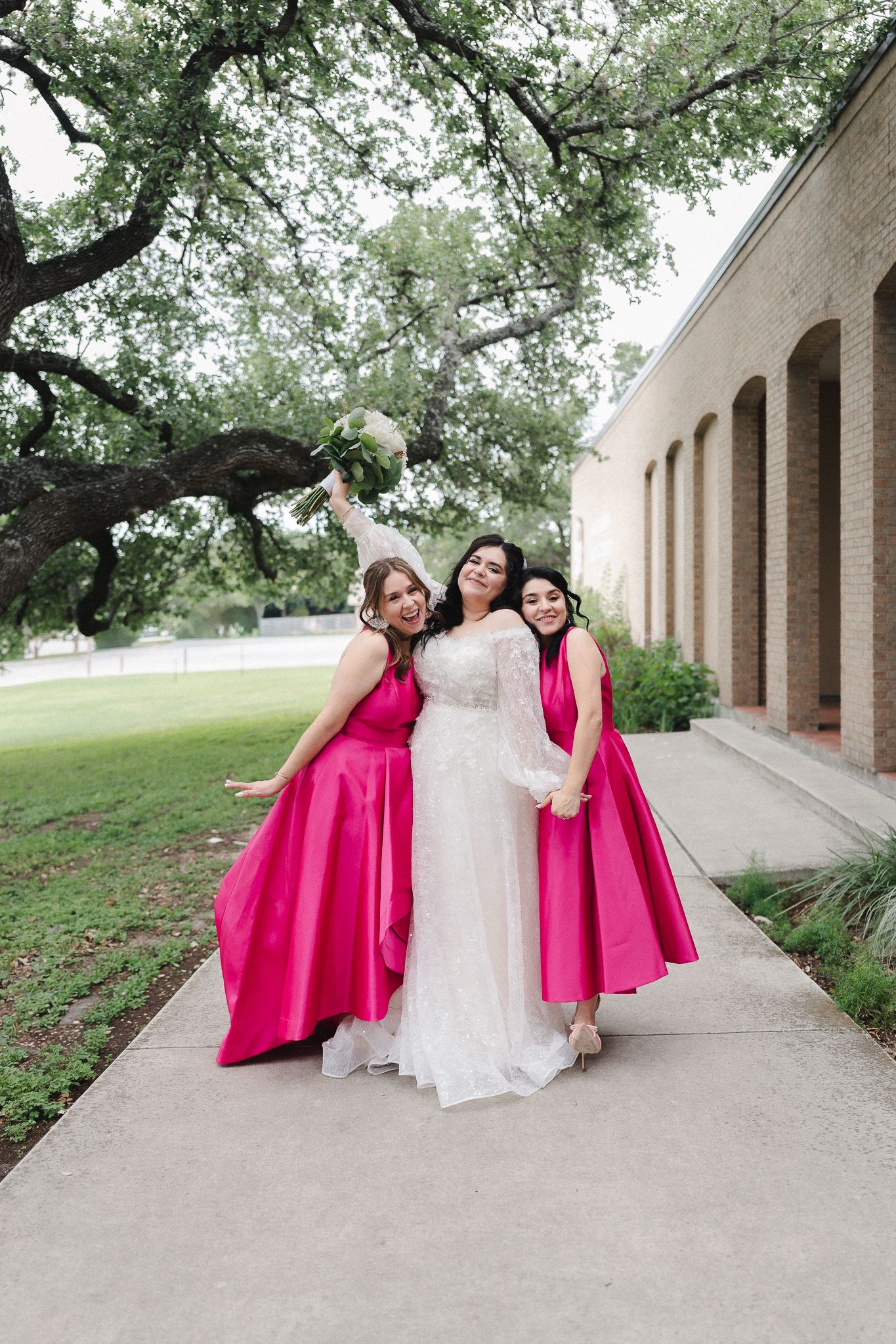 Bride in white gown poses with two bridesmaids in hot pink dresses on a pathway outside a brick building.