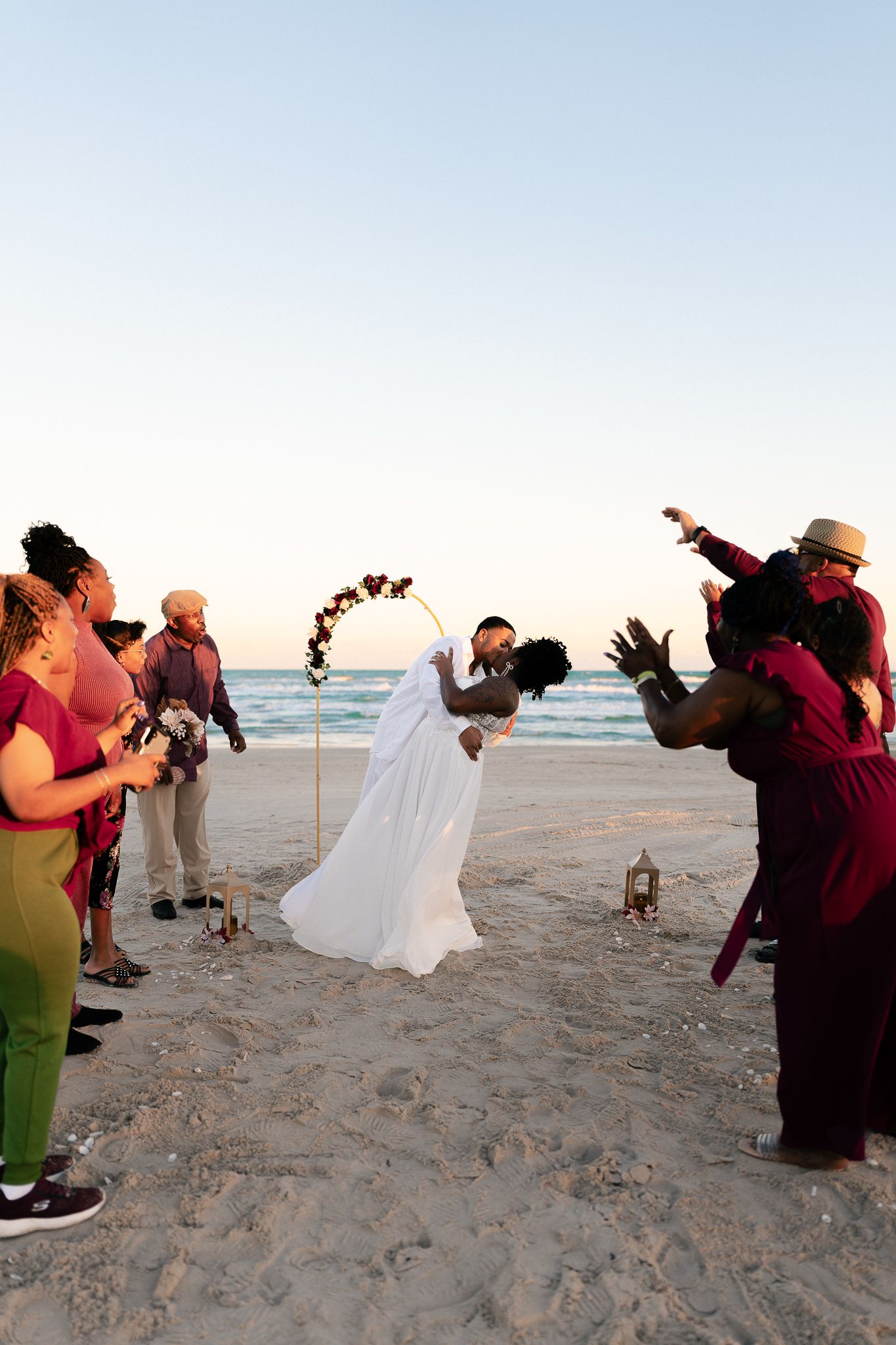 A wedding celebration on the beach at sunset with guests watching a couple dip under a floral arch.