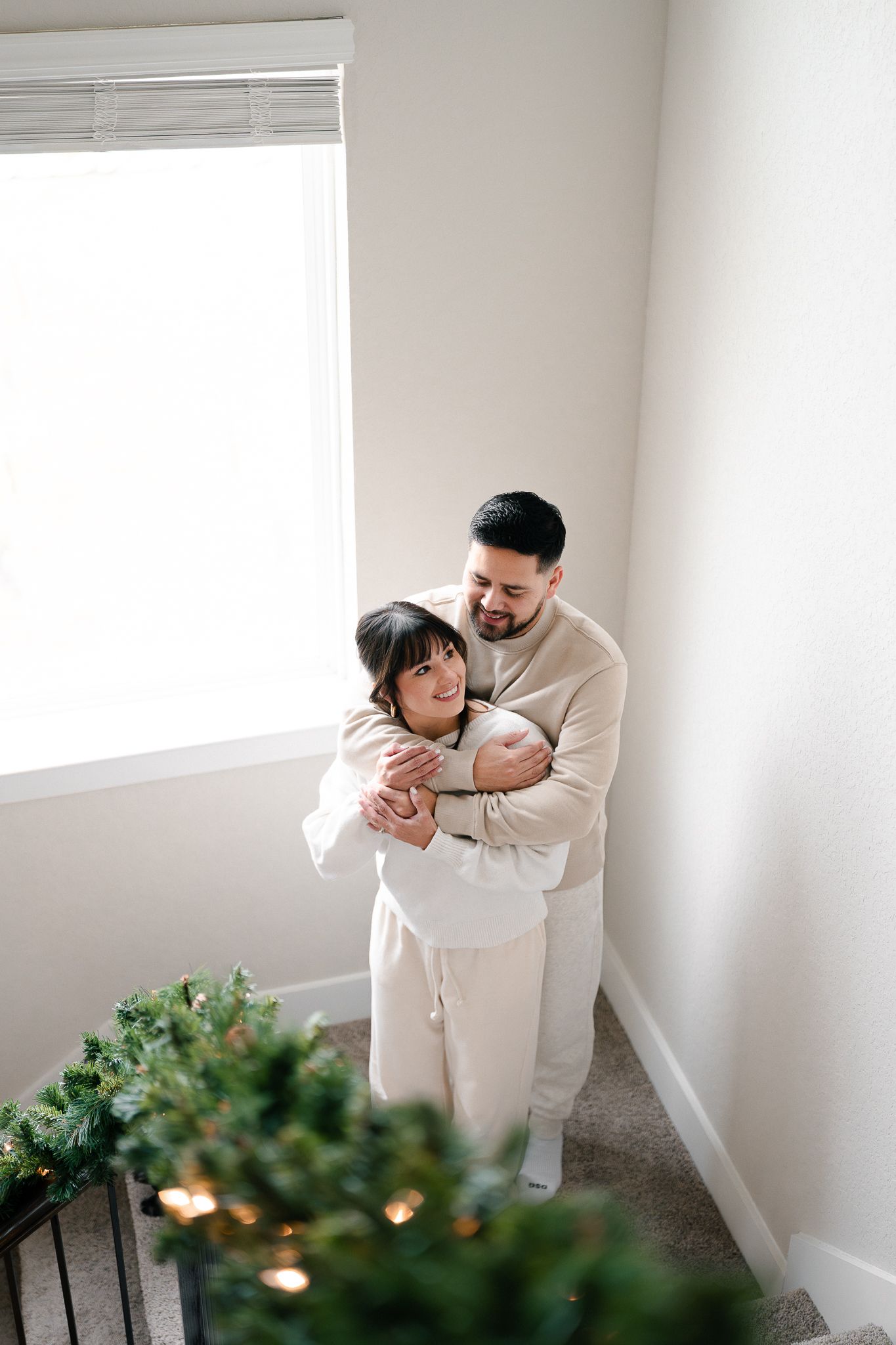 A couple embraces in a minimalist white room with green plants and twinkling lights in the foreground.