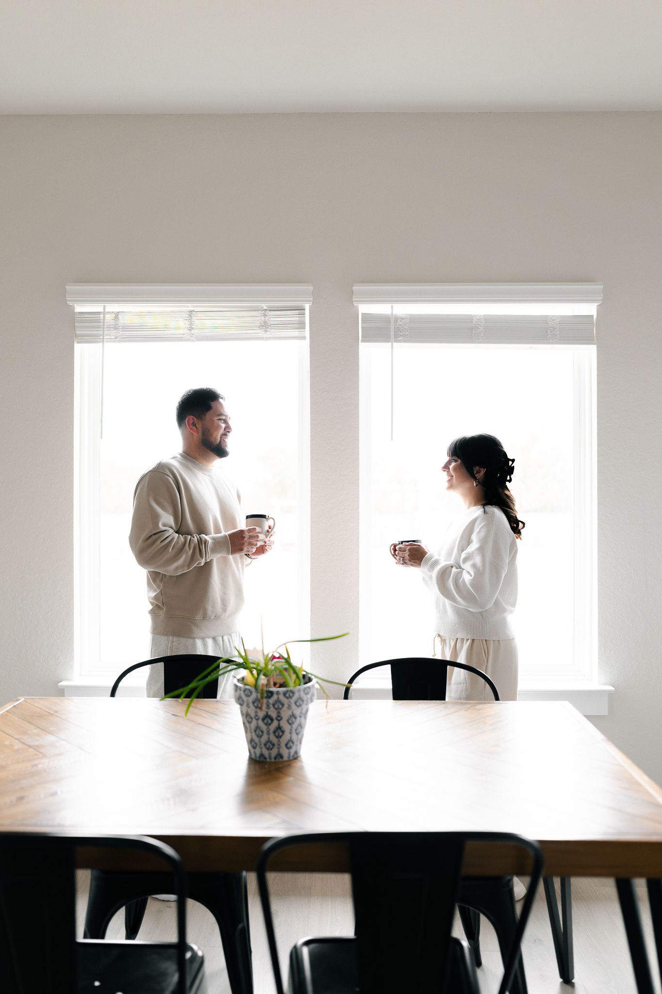 Two people in white sweaters having a conversation while standing at a long wooden table near bright windows.