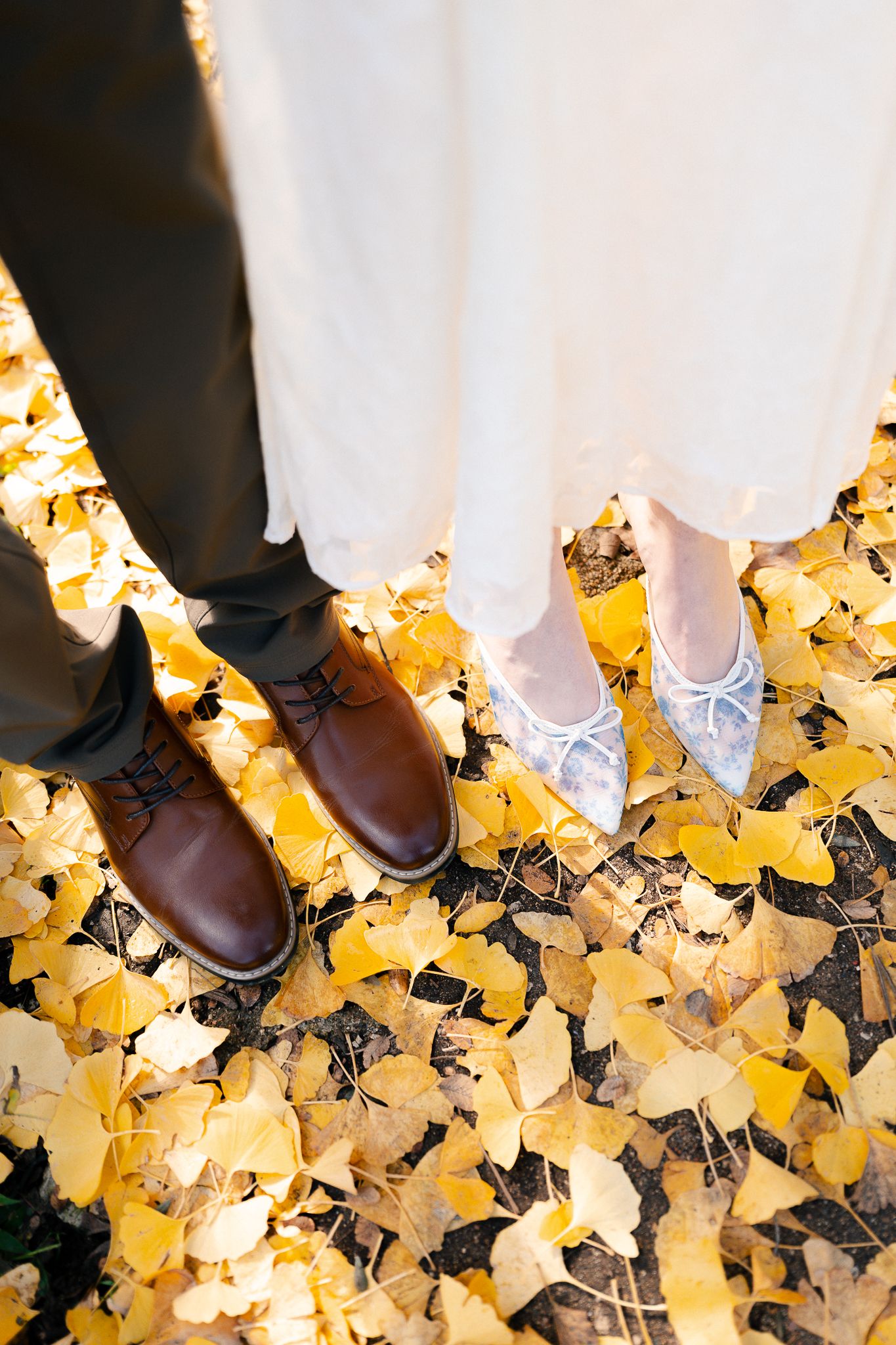 Formal footwear and dress hems photographed on carpet of bright yellow fall leaves.