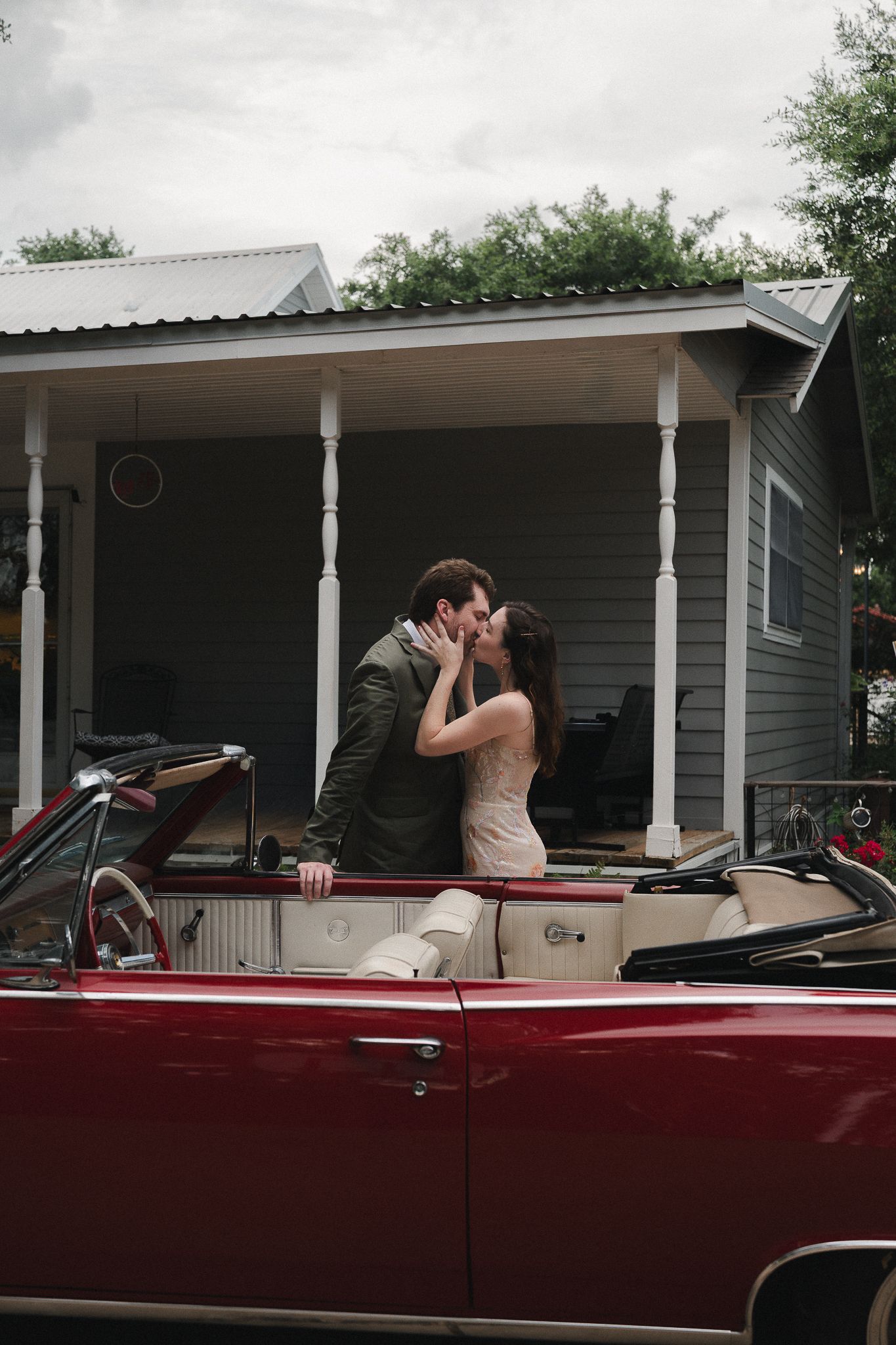 Couple kissing while standing in a red vintage convertible car in front of a charming house with a covered porch.