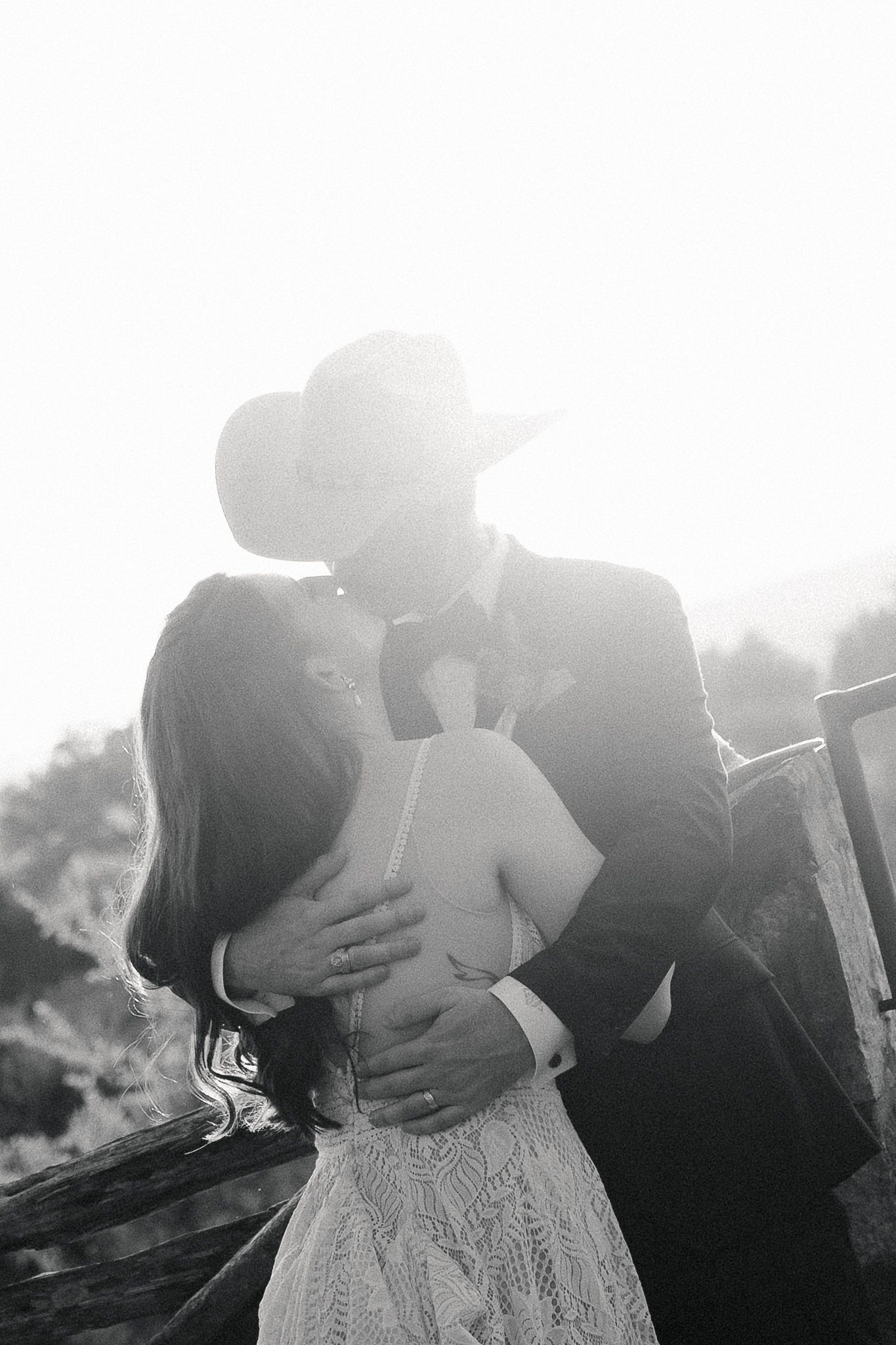 Black and white photo of a couple sharing a romantic kiss, groom in cowboy hat and suit embracing bride in lace dress.