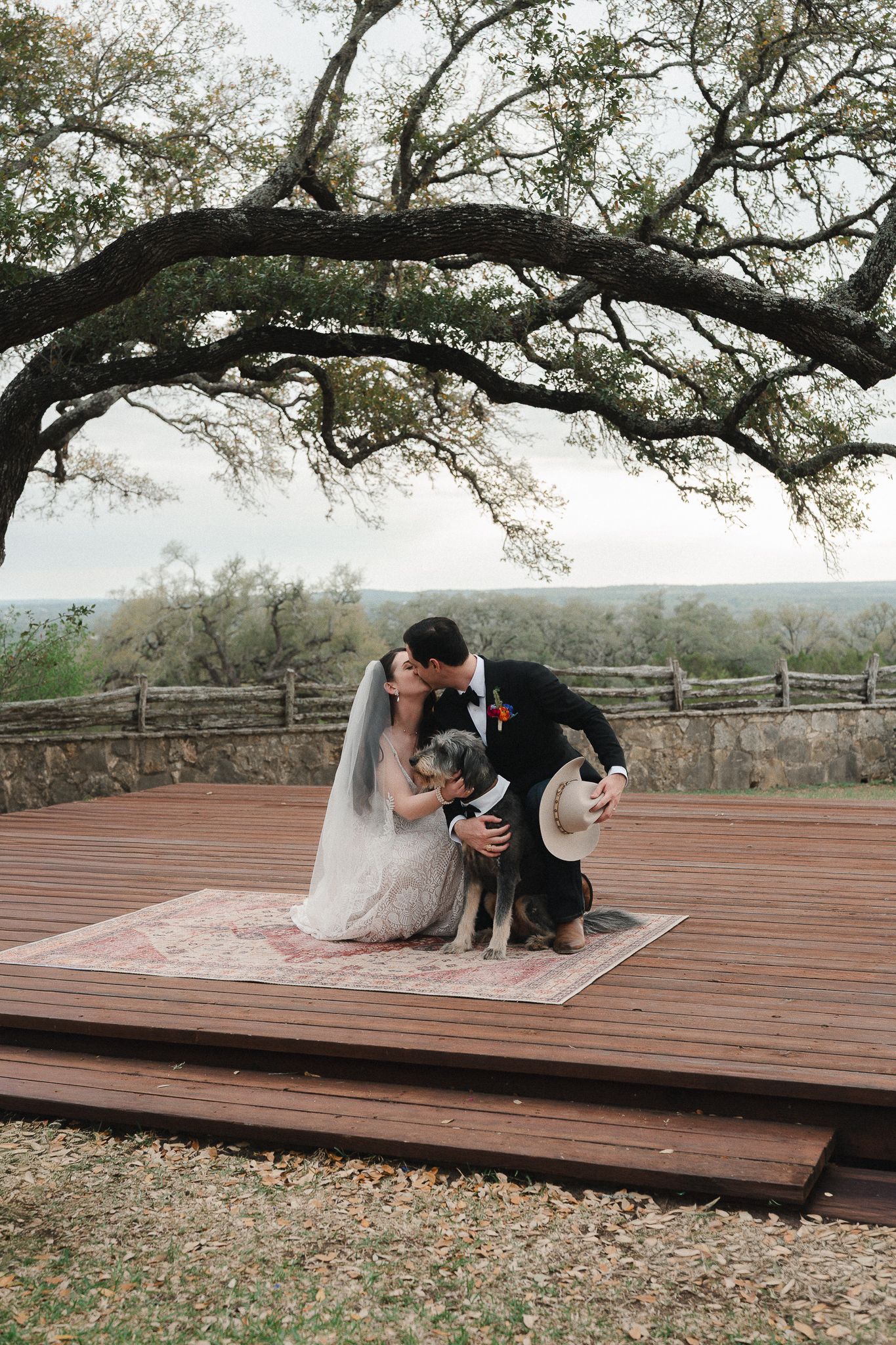 Bride and groom kissing on rustic wooden deck with their dog, framed by oak trees and scenic Texas Hill Country views.
