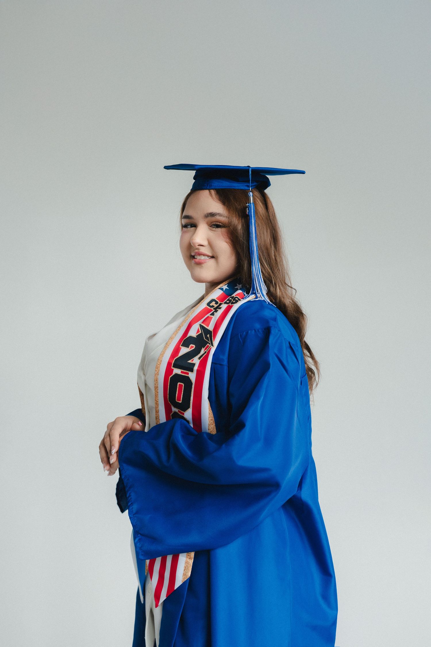 Young woman in blue graduation cap and gown with sash, smiling against a light gray background.