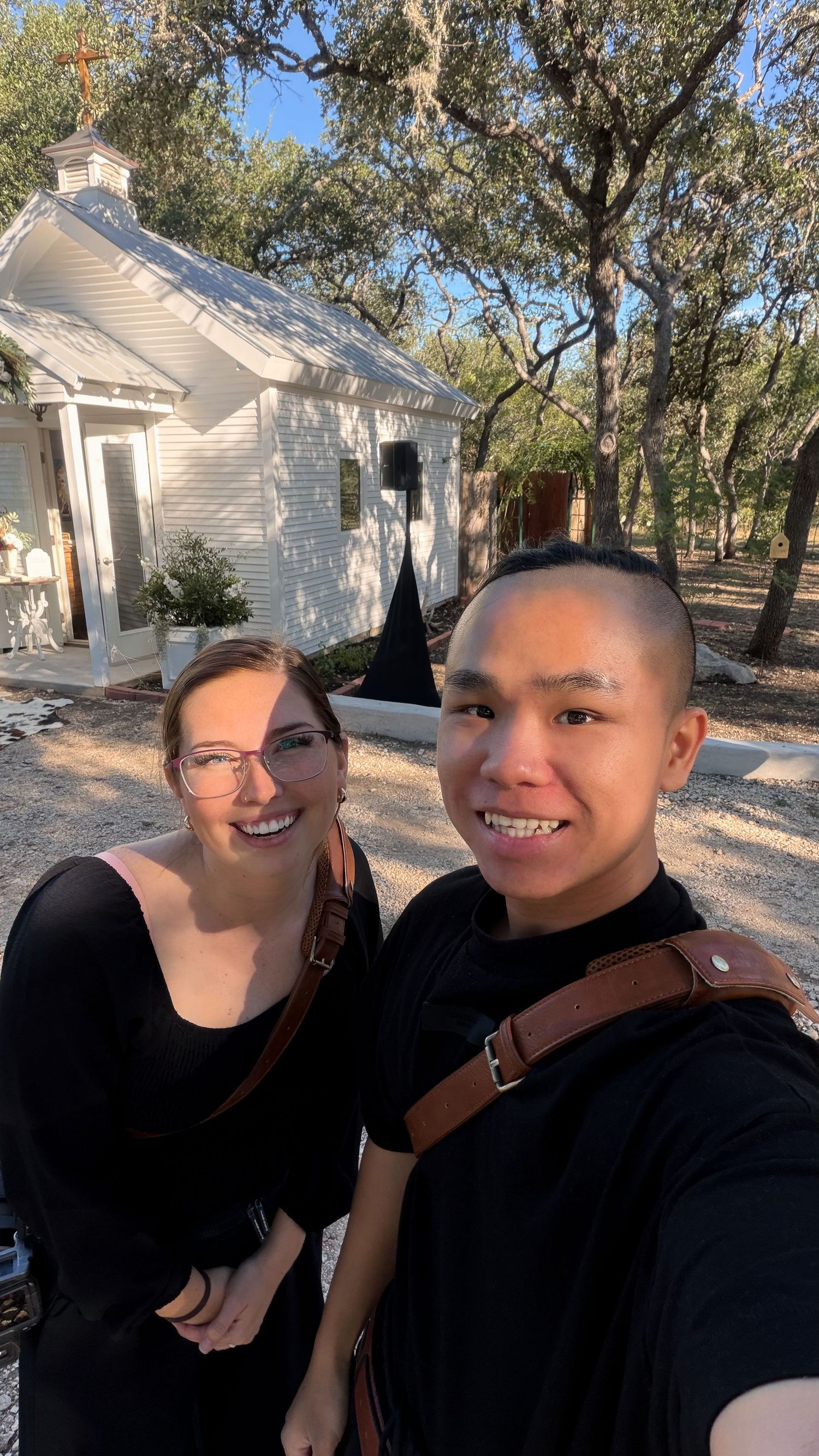 Two smiling people in black outfits with leather camera straps take a selfie in front of a small white chapel surrounded by oak trees.