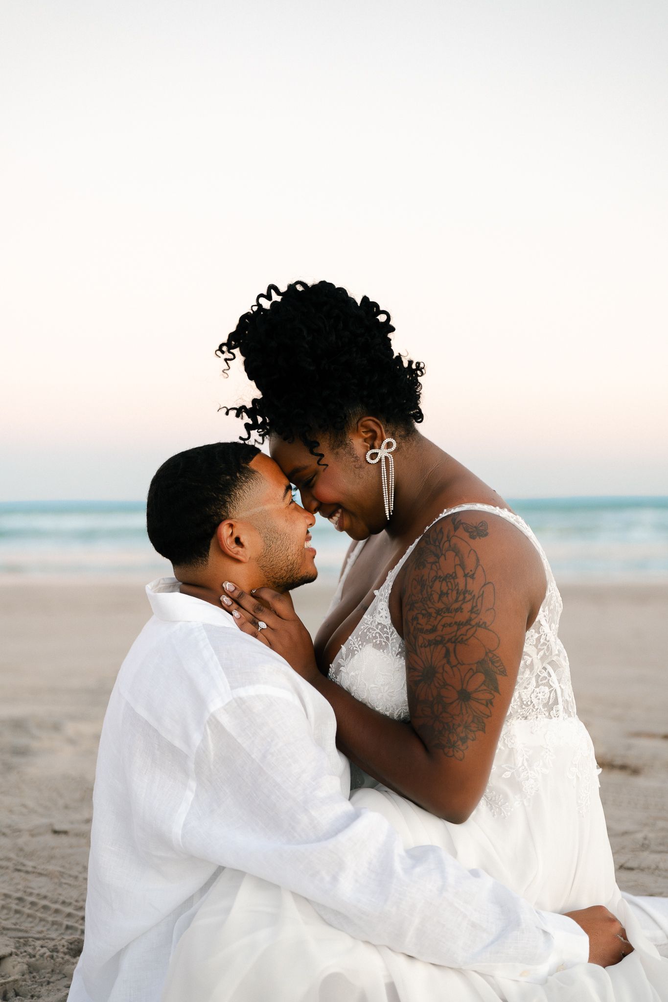 A loving couple shares intimate moments on a serene beach at sunset wearing matching white outfits.