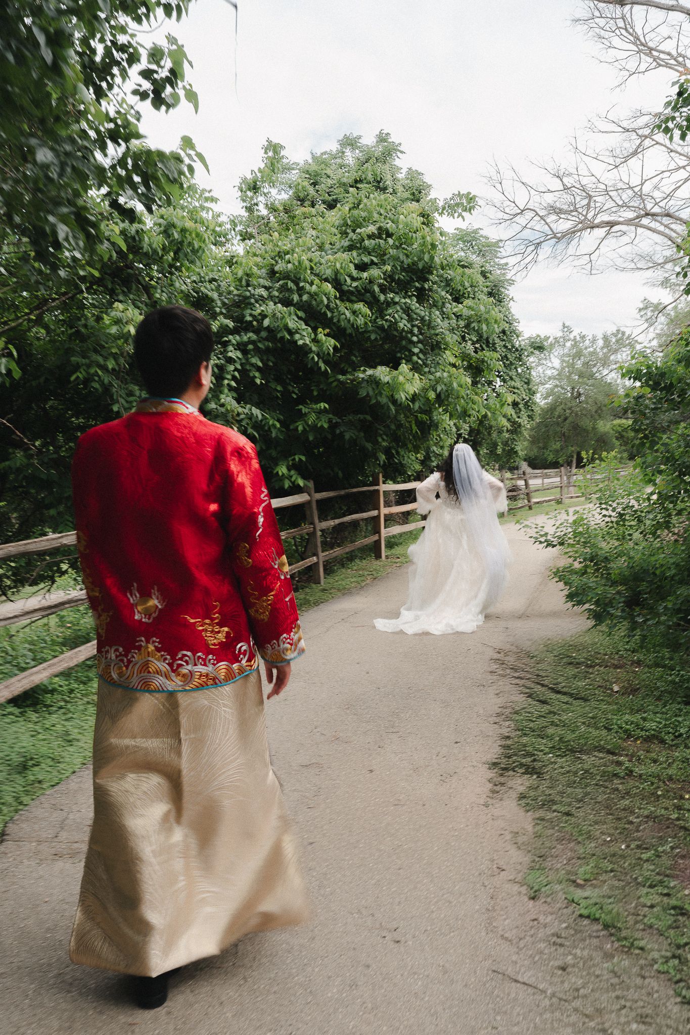 Couple in traditional Chinese wedding attire walking on a garden path, bride in white gown with veil, groom in red embroidered jacket.