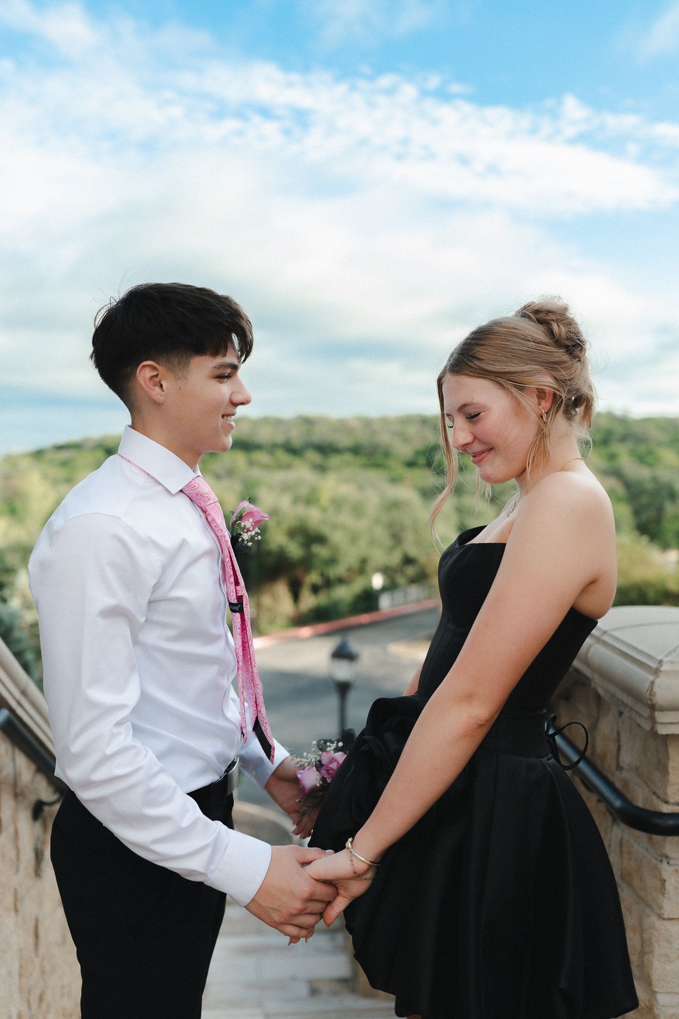 A young couple in formal attire holds hands and smiles at each other outdoors under a blue sky.