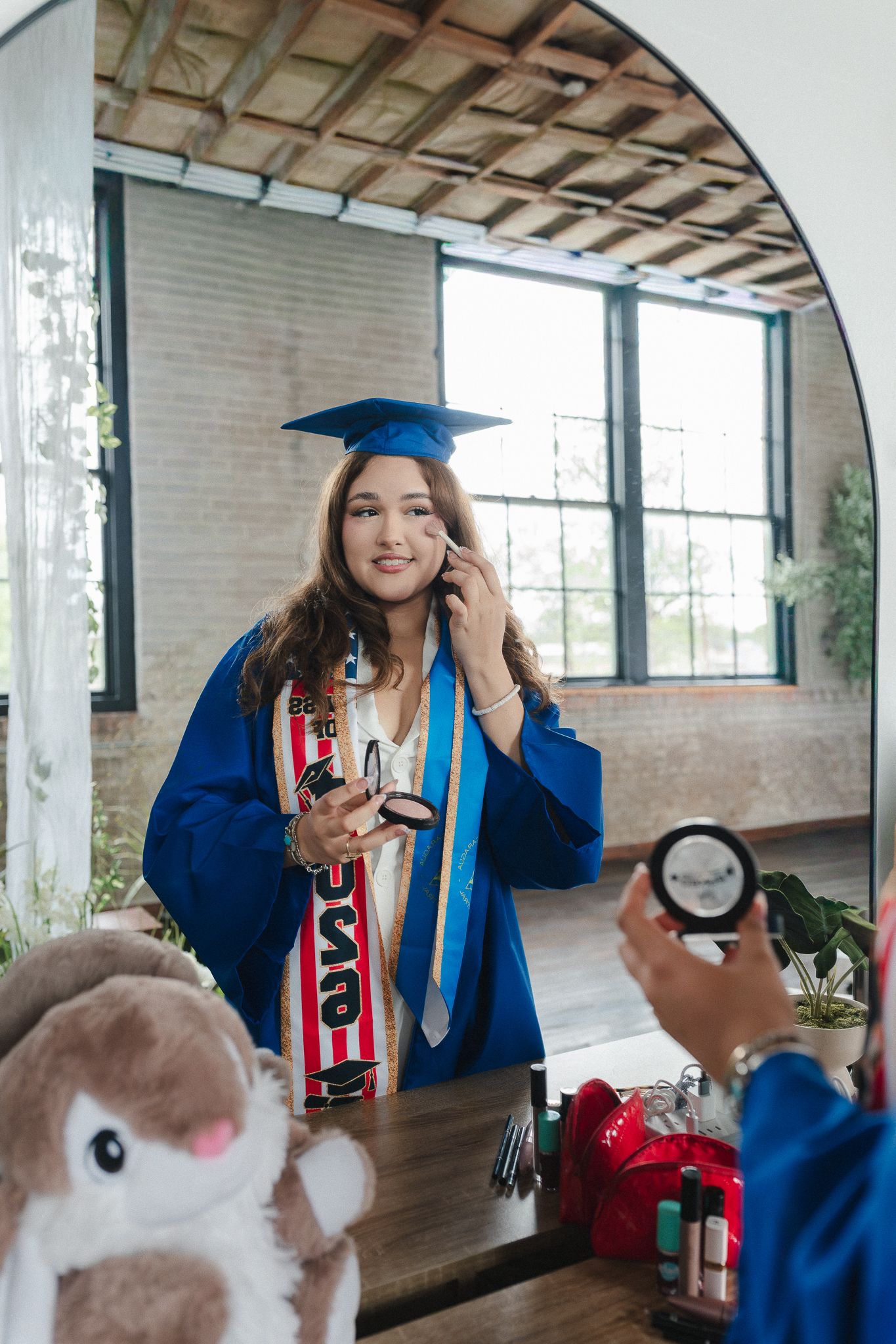 A young woman in blue graduation cap and gown applies makeup while looking in an arched mirror.