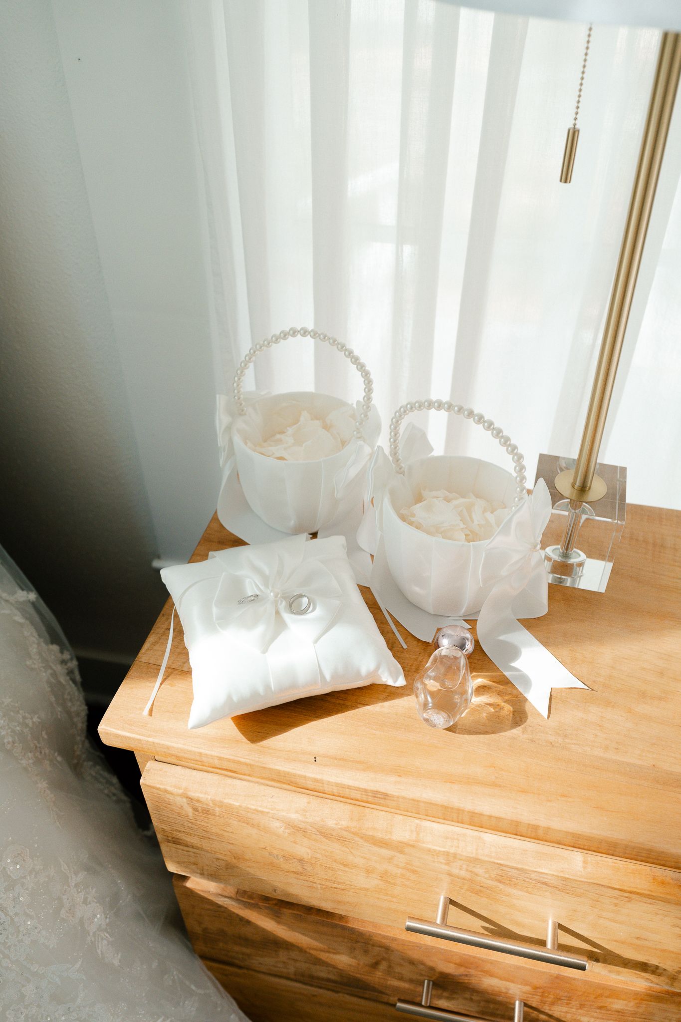 Wedding accessories including white flower baskets and ring pillow displayed on wooden dresser near sheer curtains.