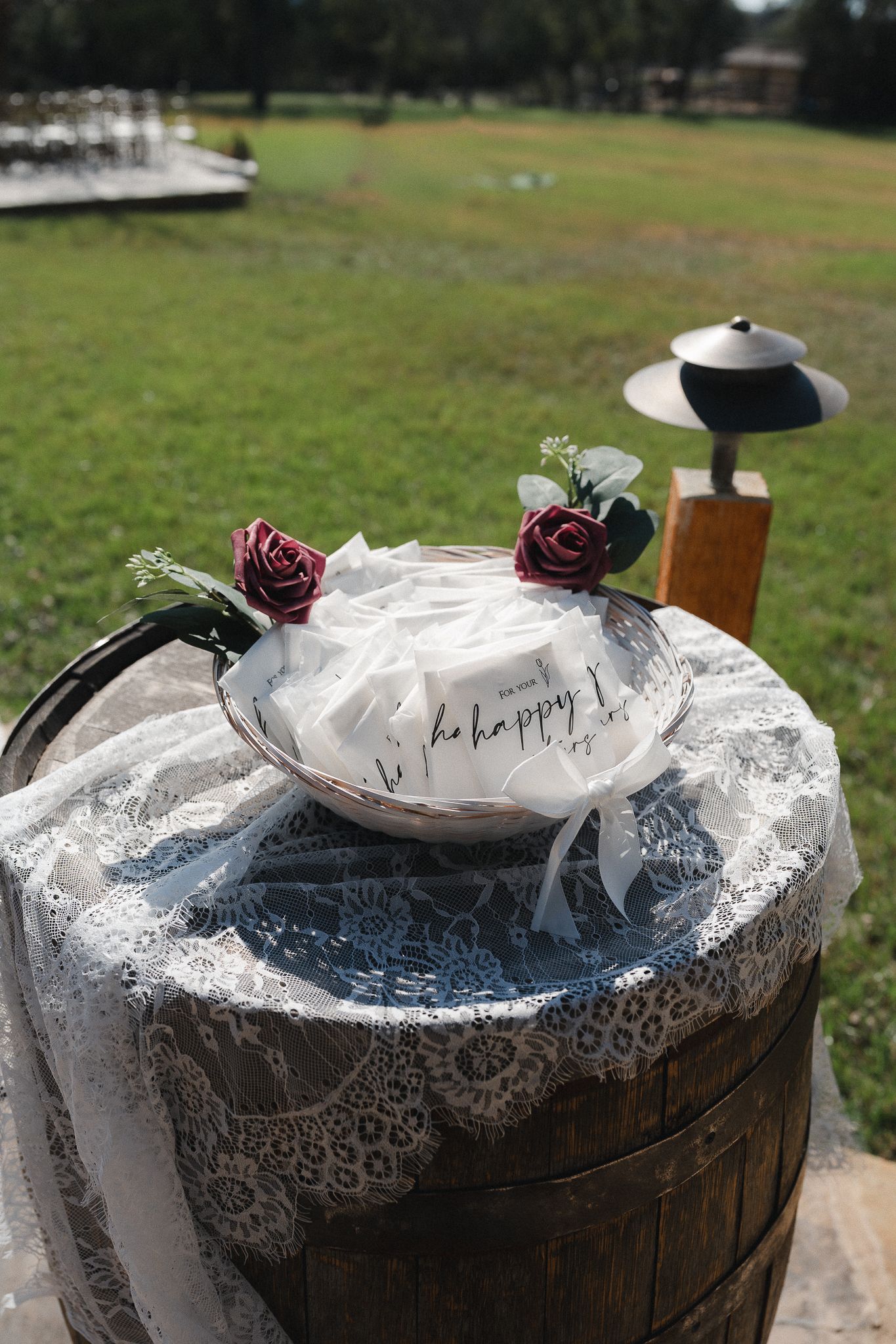 Rustic wedding favor display on a wooden barrel with lace, white bags, and burgundy roses outdoors.
