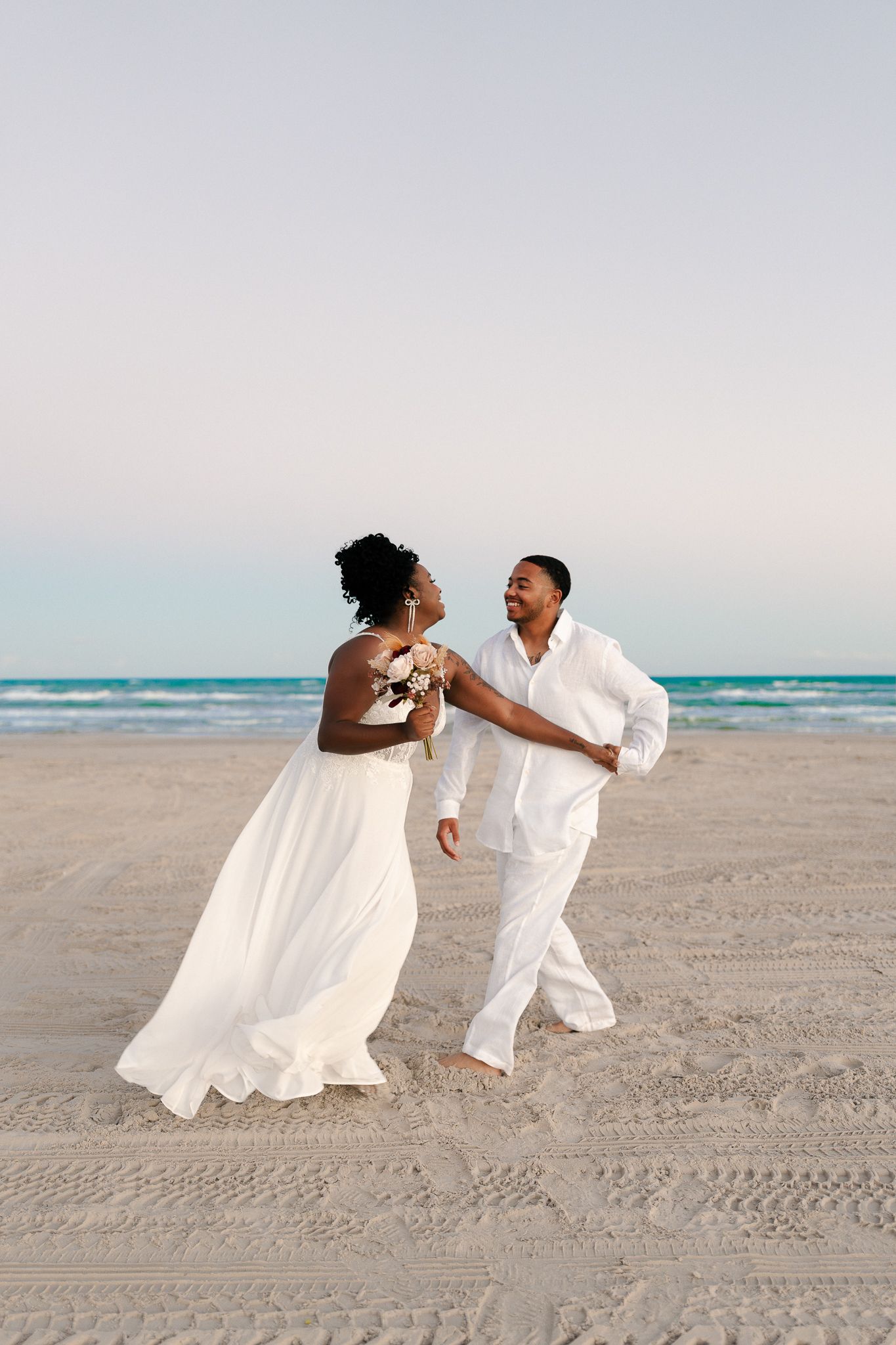 A couple in white attire dances joyfully together on a beach at sunset with ocean waves in the background.