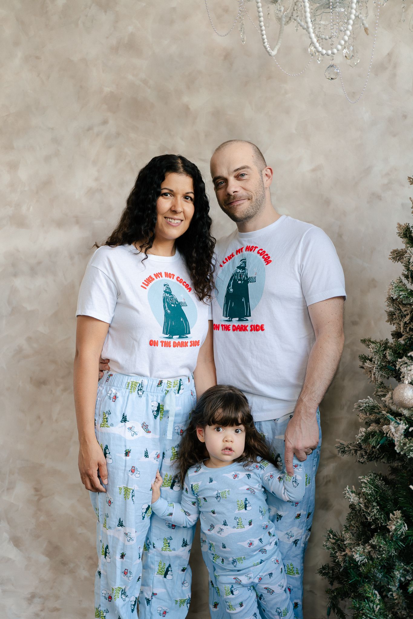 Family wearing matching dinosaur print pajama pants and t-shirts pose together against textured wall.