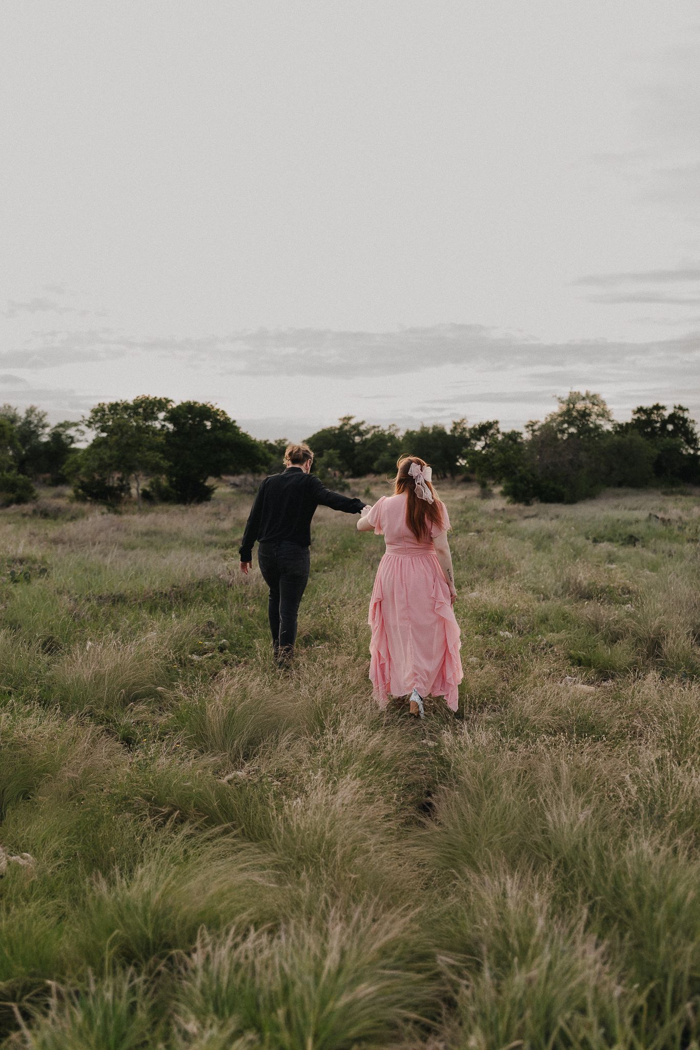 A couple walks hand-in-hand through a lush green meadow, woman in pink dress, man in dark suit, trees in background.