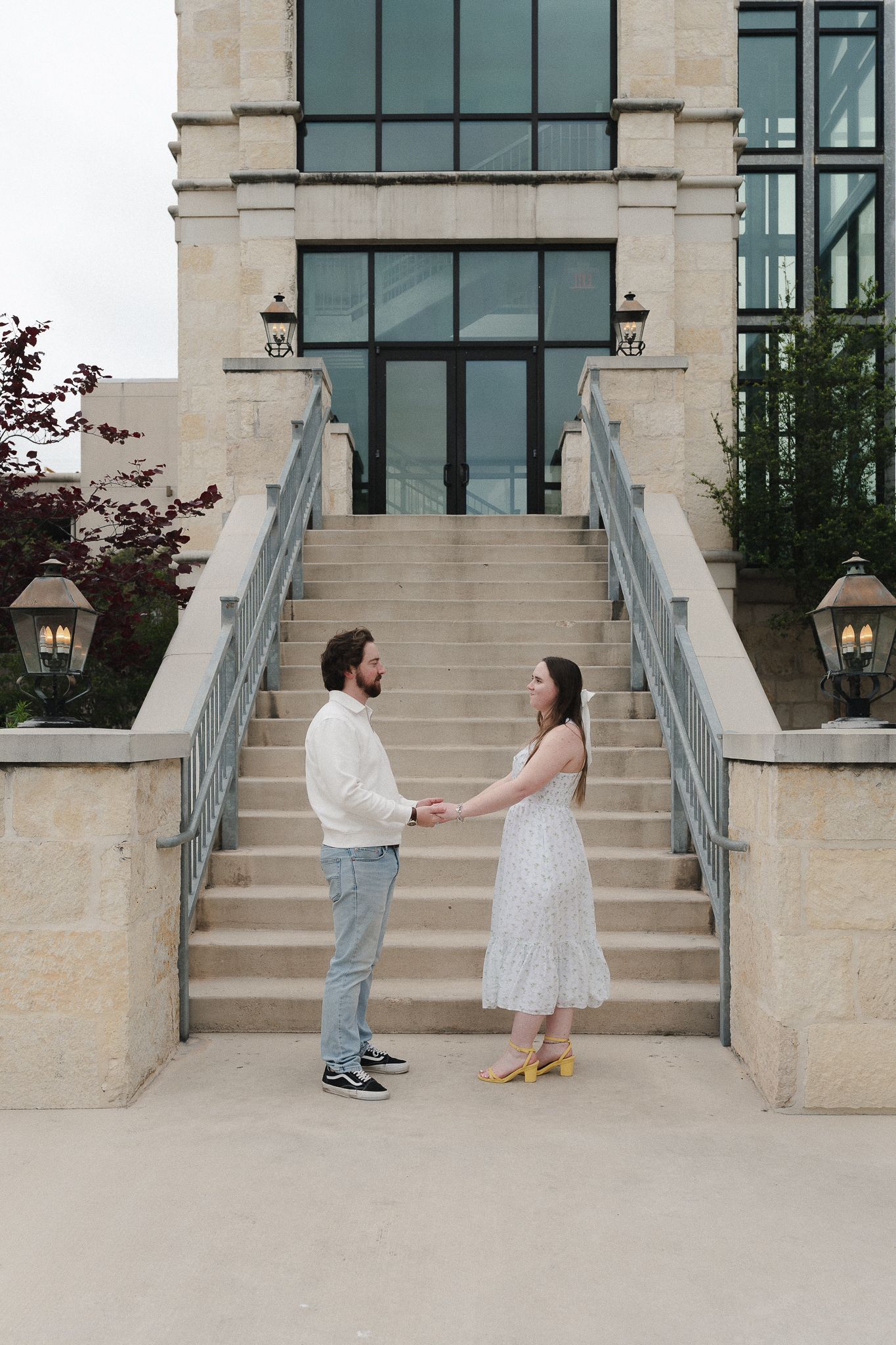 Couple holds hands facing each other on stone steps of elegant building with glass windows and lanterns.