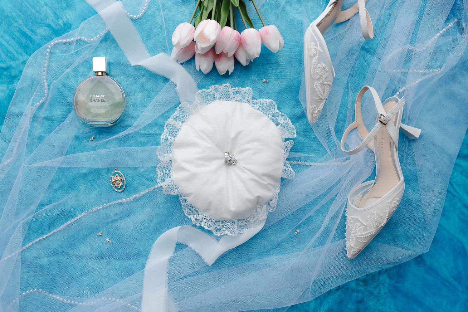 Elegant wedding flat lay with lace heels, ring pillow, pink tulips, perfume, and veil on blue background.