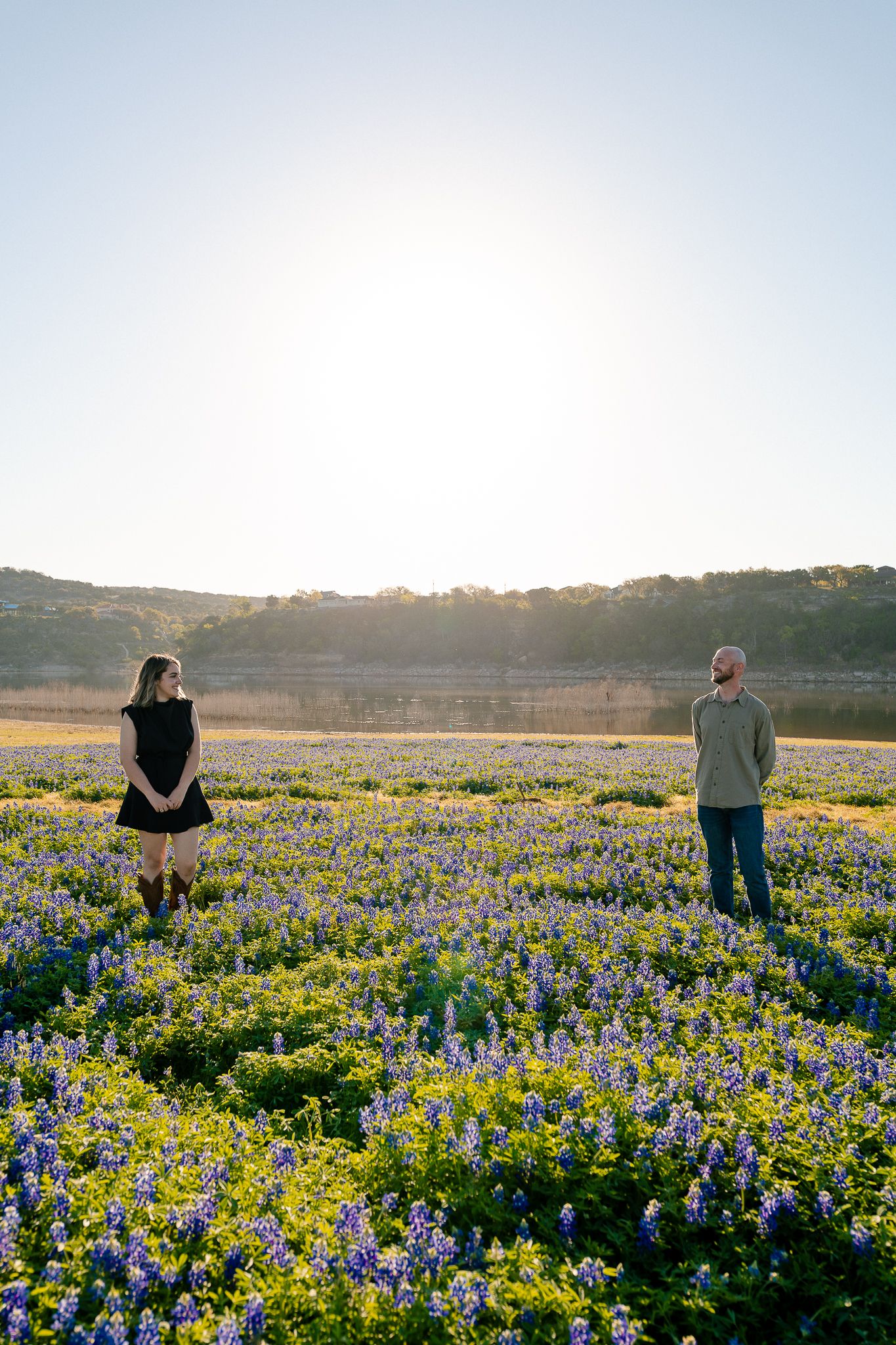 Two people standing apart in a vast Texas bluebonnet field at golden hour with rolling hills in the background.