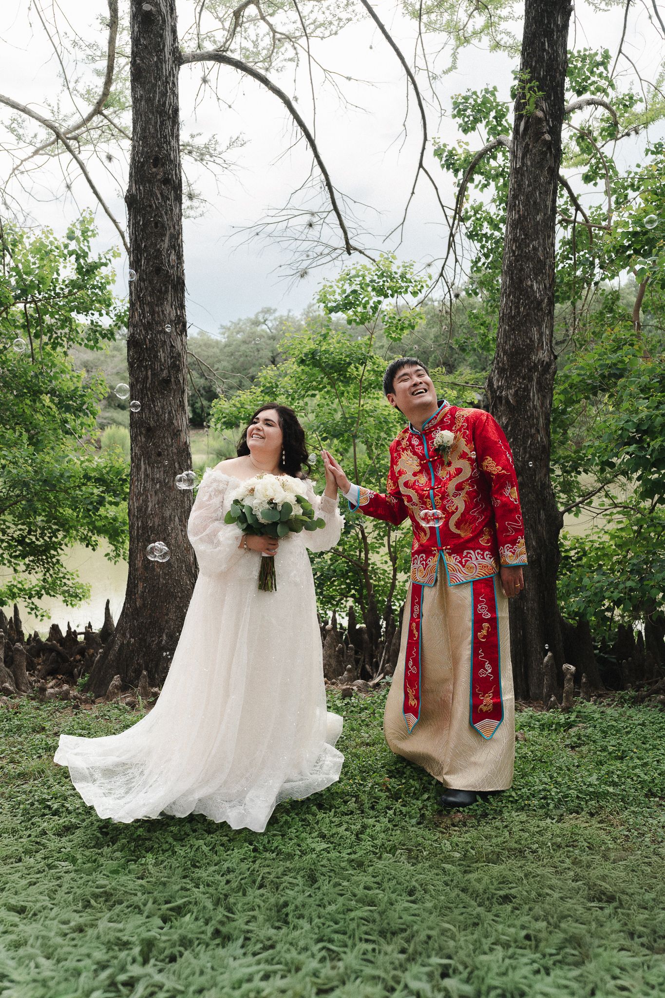 Bride in white lace gown and groom in red Chinese traditional attire laugh while playing with bubbles outdoors.