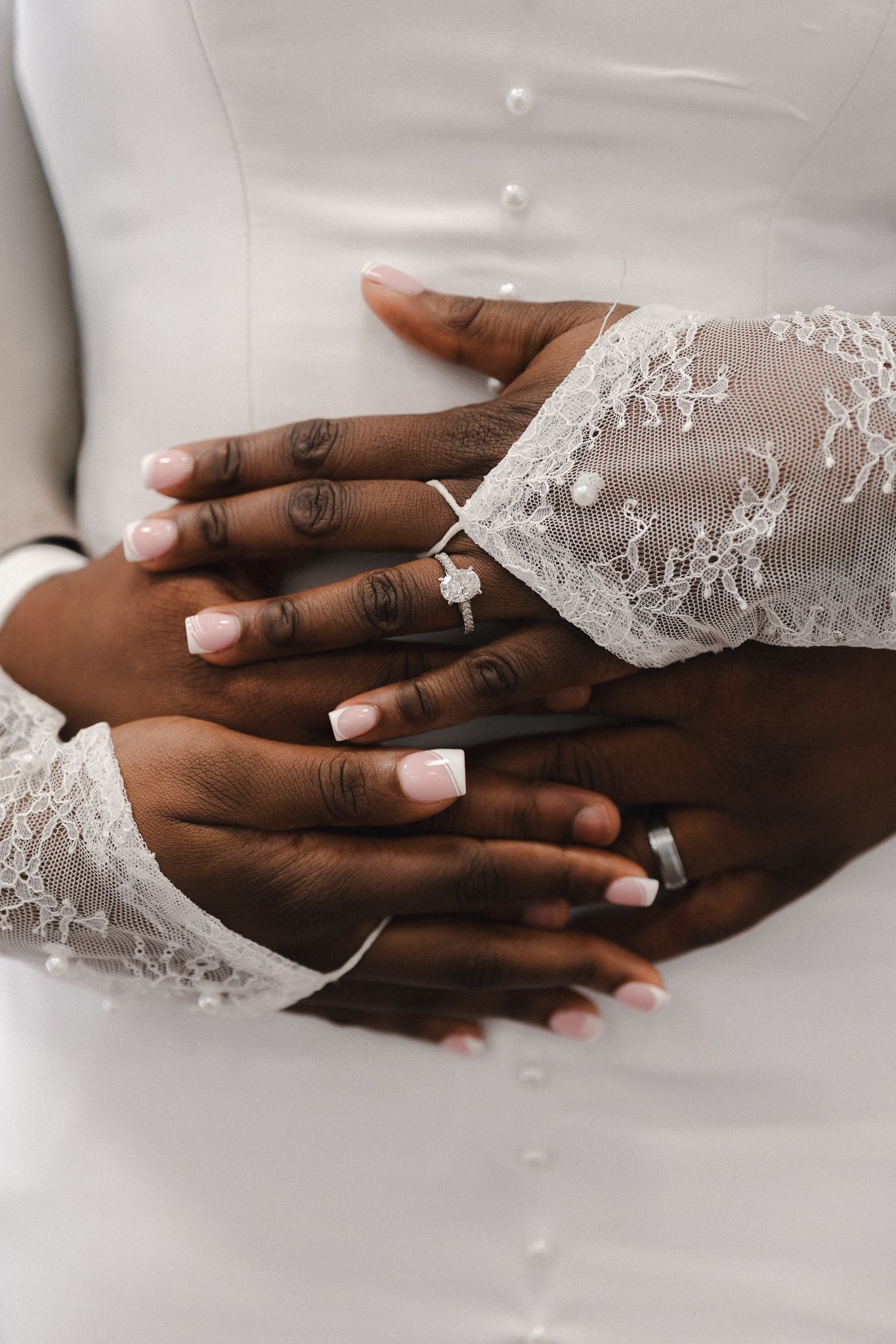 Close-up of newlywed couple's hands showing diamond engagement ring and wedding band on white lace bridal gown.