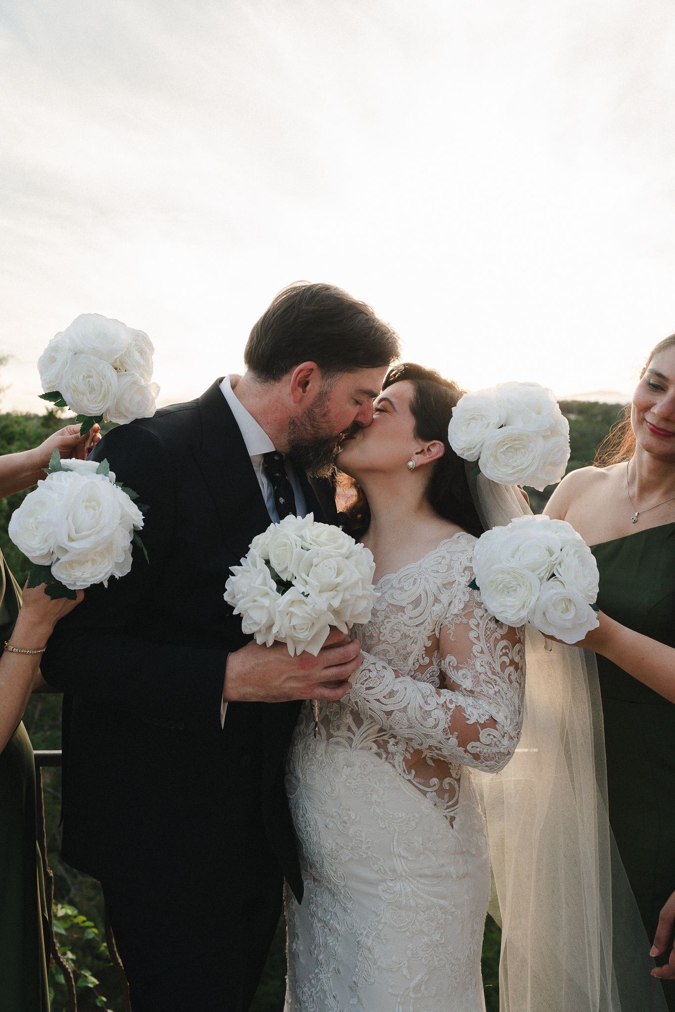 Bride in a lace wedding gown and groom in a dark suit kiss at sunset, surrounded by bridesmaids holding white rose bouquets.