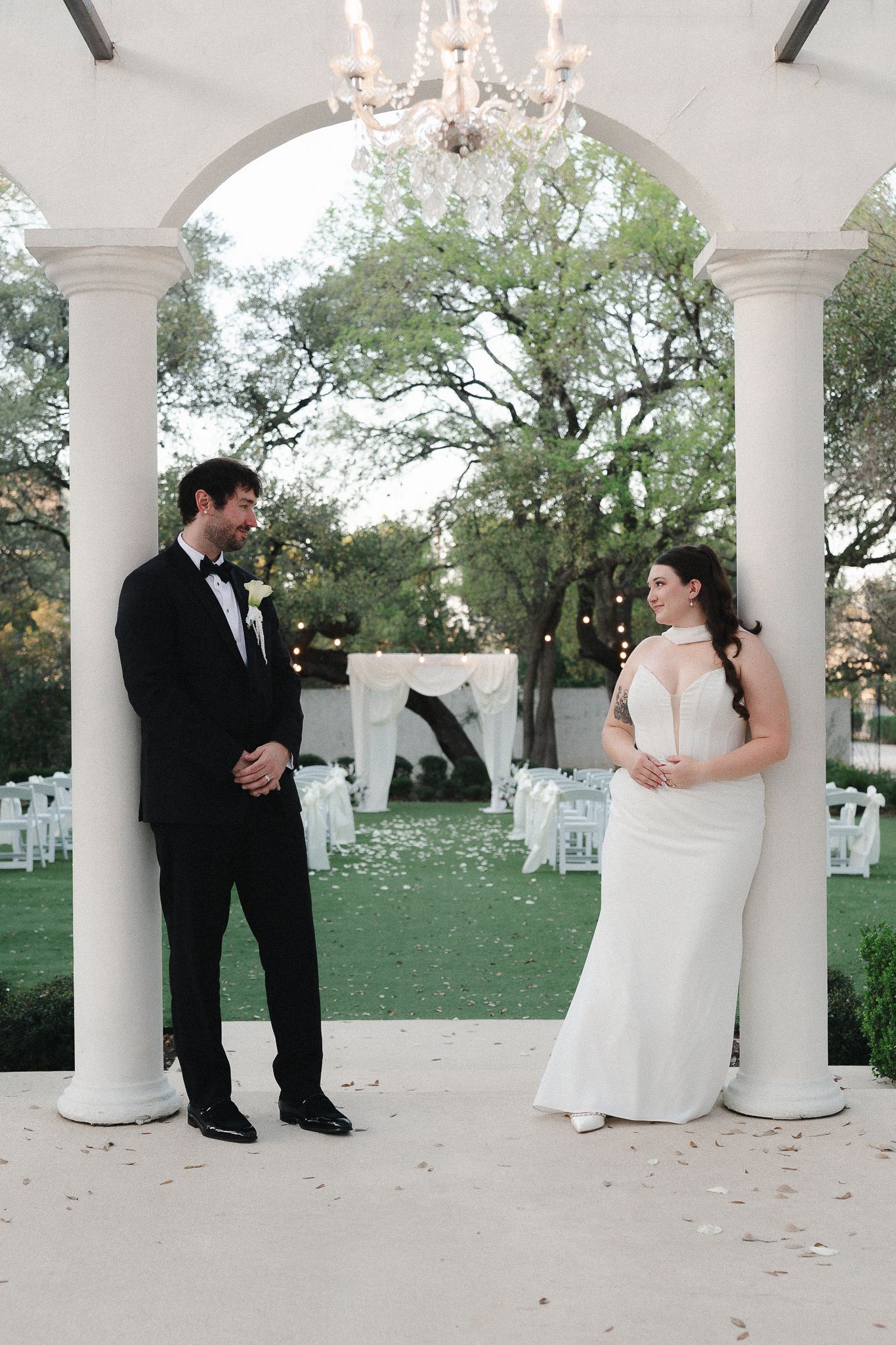Bride and groom share a joyful first look under a white columned pavilion with chandelier at an elegant outdoor wedding venue.