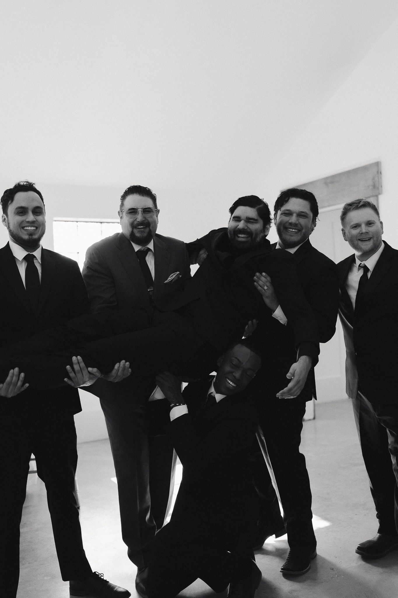 Black and white photo of six men in suits laughing and posing together, one being held horizontally by the group.