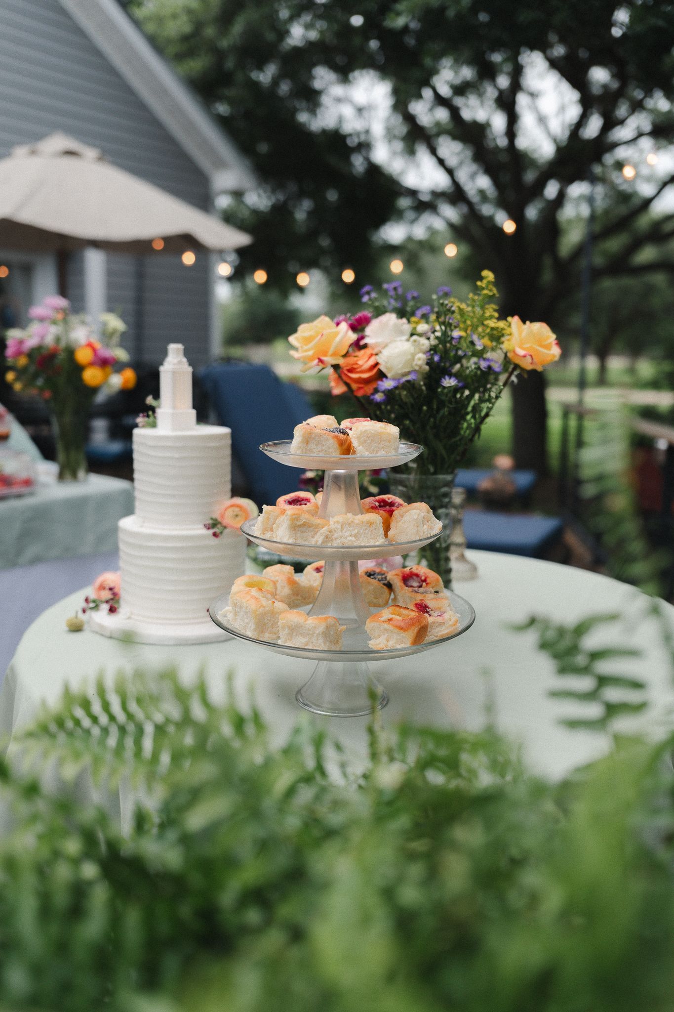 Elegant outdoor wedding dessert table with white tiered cake, cupcake stand, and colorful floral arrangements in a garden setting.