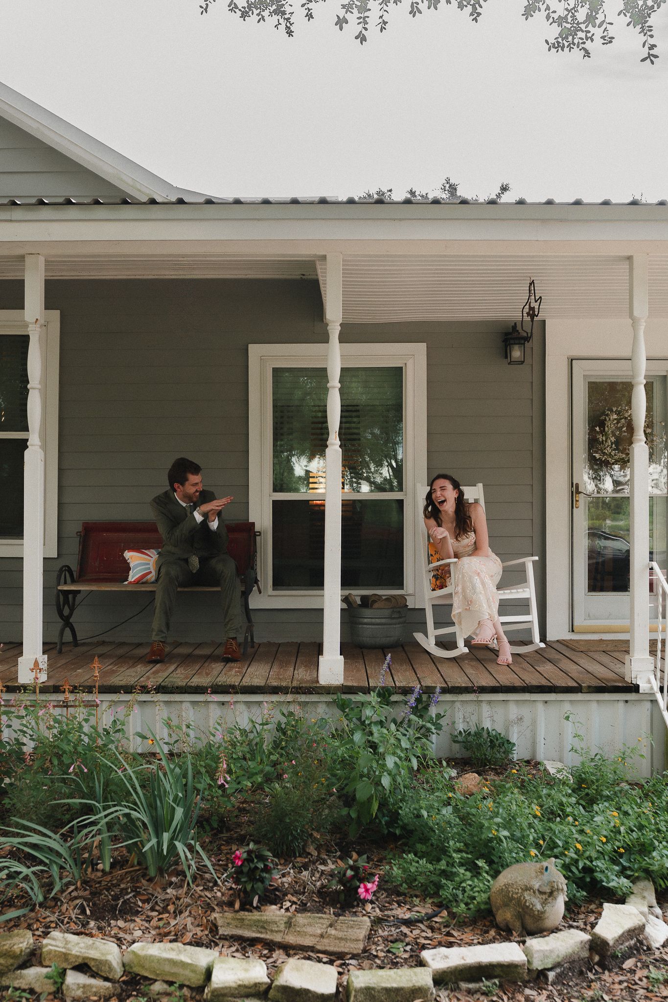 Couple relaxing on a charming farmhouse porch, man sitting on bench and woman in white rocking chair, surrounded by lush garden.