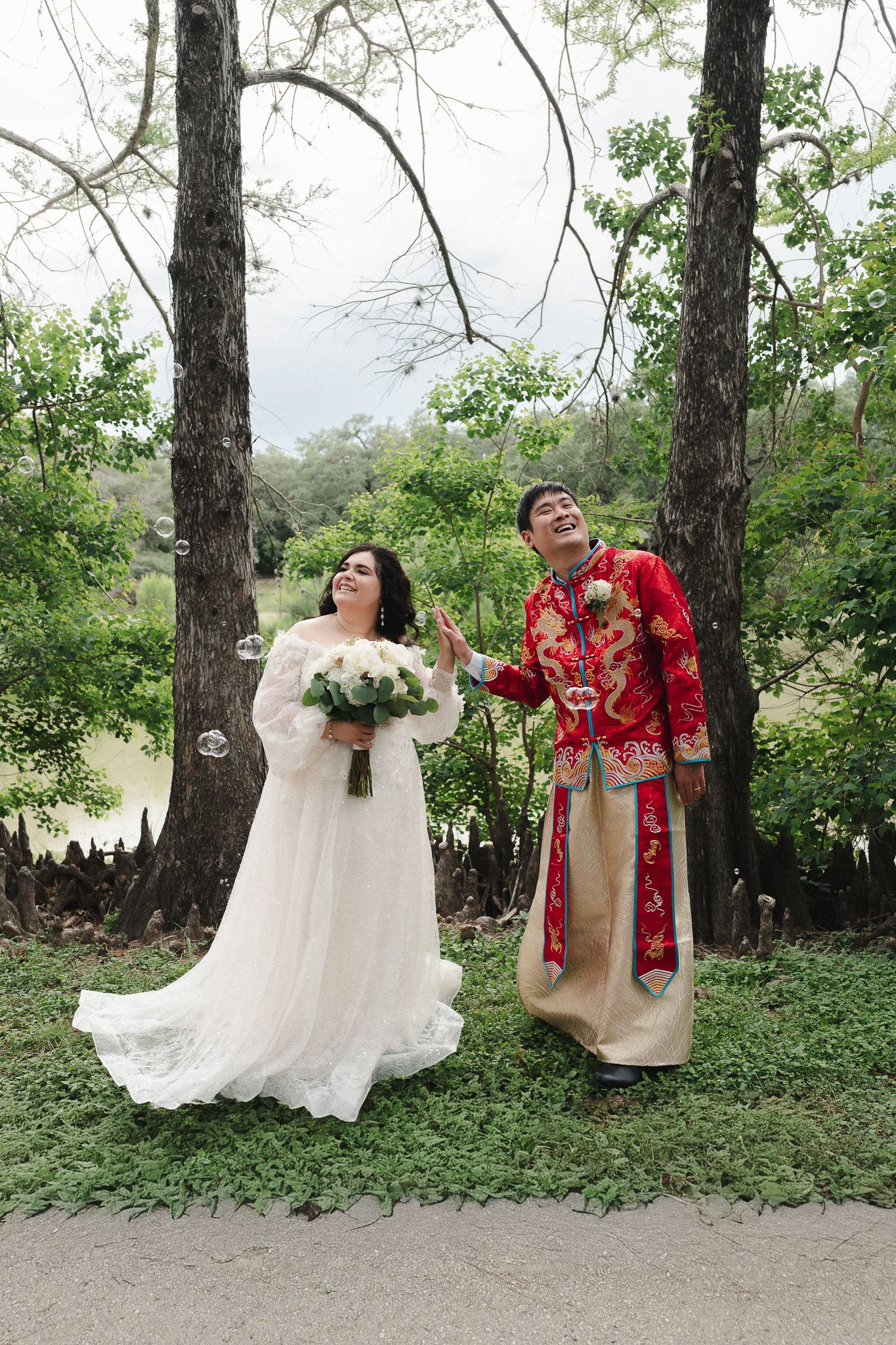 Couple in wedding attire, bride in white gown and groom in red Chinese suit, posing outdoors among green trees.