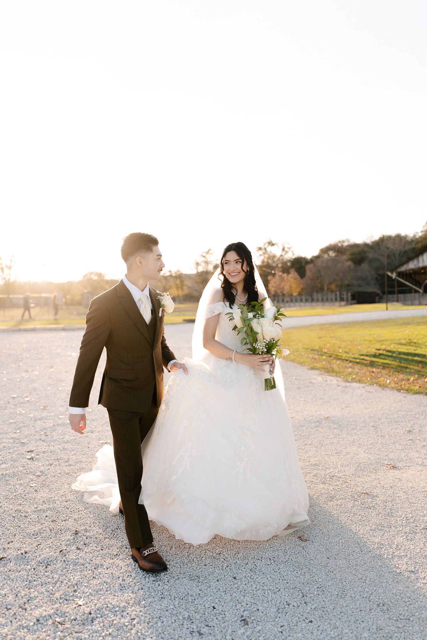A couple in wedding attire walking together on a gravel path during golden hour sunset.