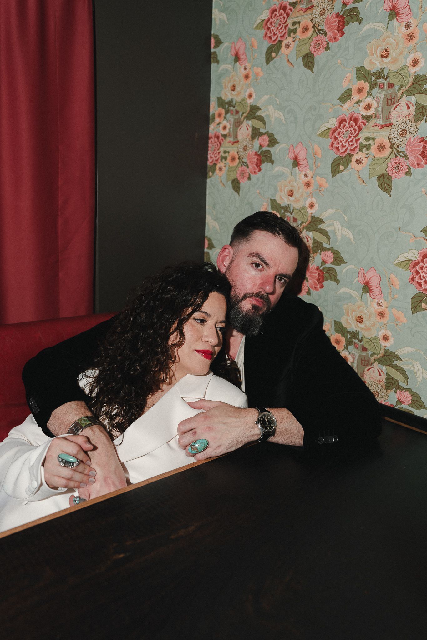A couple poses intimately at a dark table, woman in white suit with turquoise rings, man in black jacket, floral wallpaper behind.