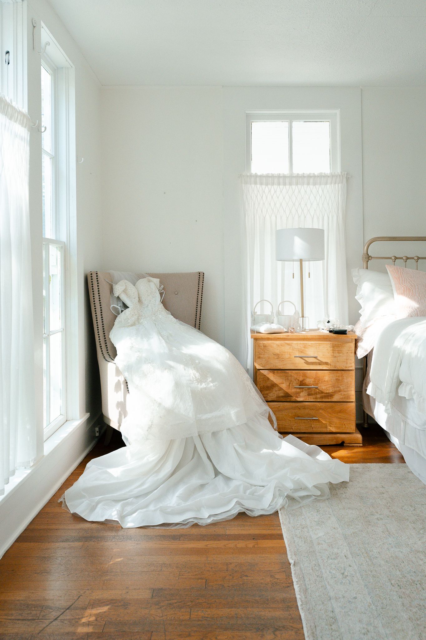 White wedding dress draped over a chair in a bright bedroom with wood floors and rustic dresser.