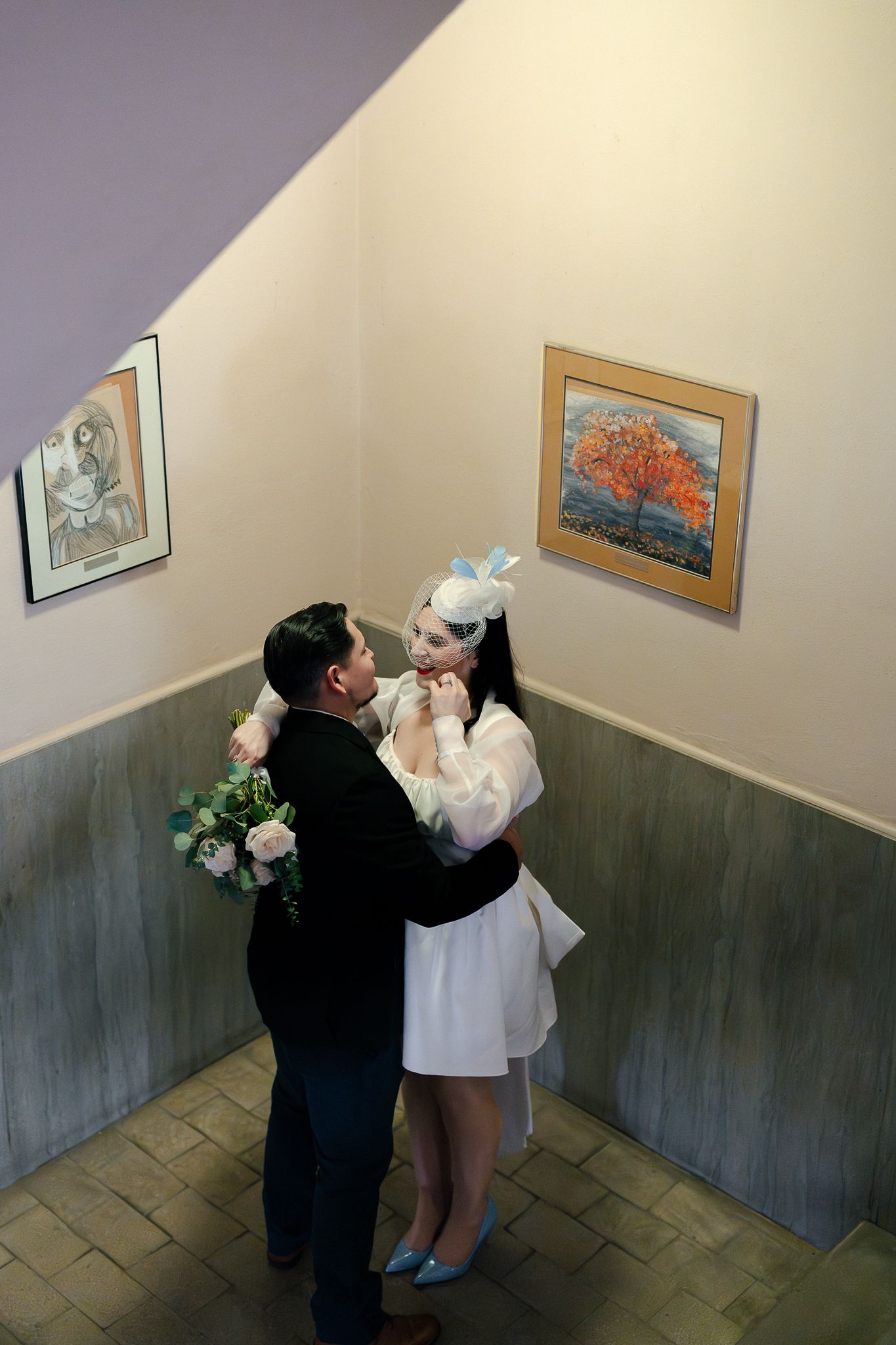 Newlywed couple embracing in an art gallery, bride in white dress with floral headpiece holding bouquet.