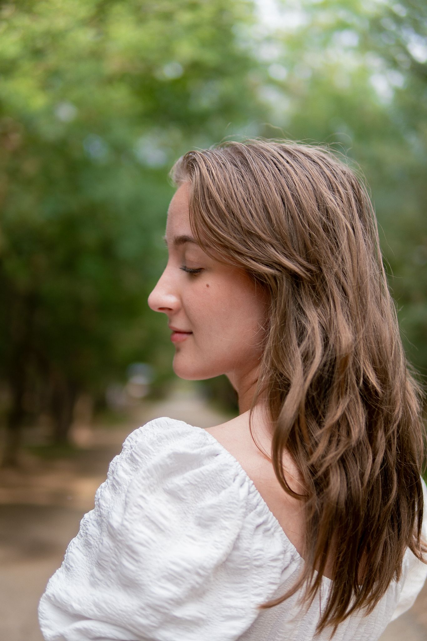 Side profile portrait of someone with long wavy brown hair wearing a white blouse in an outdoor setting.