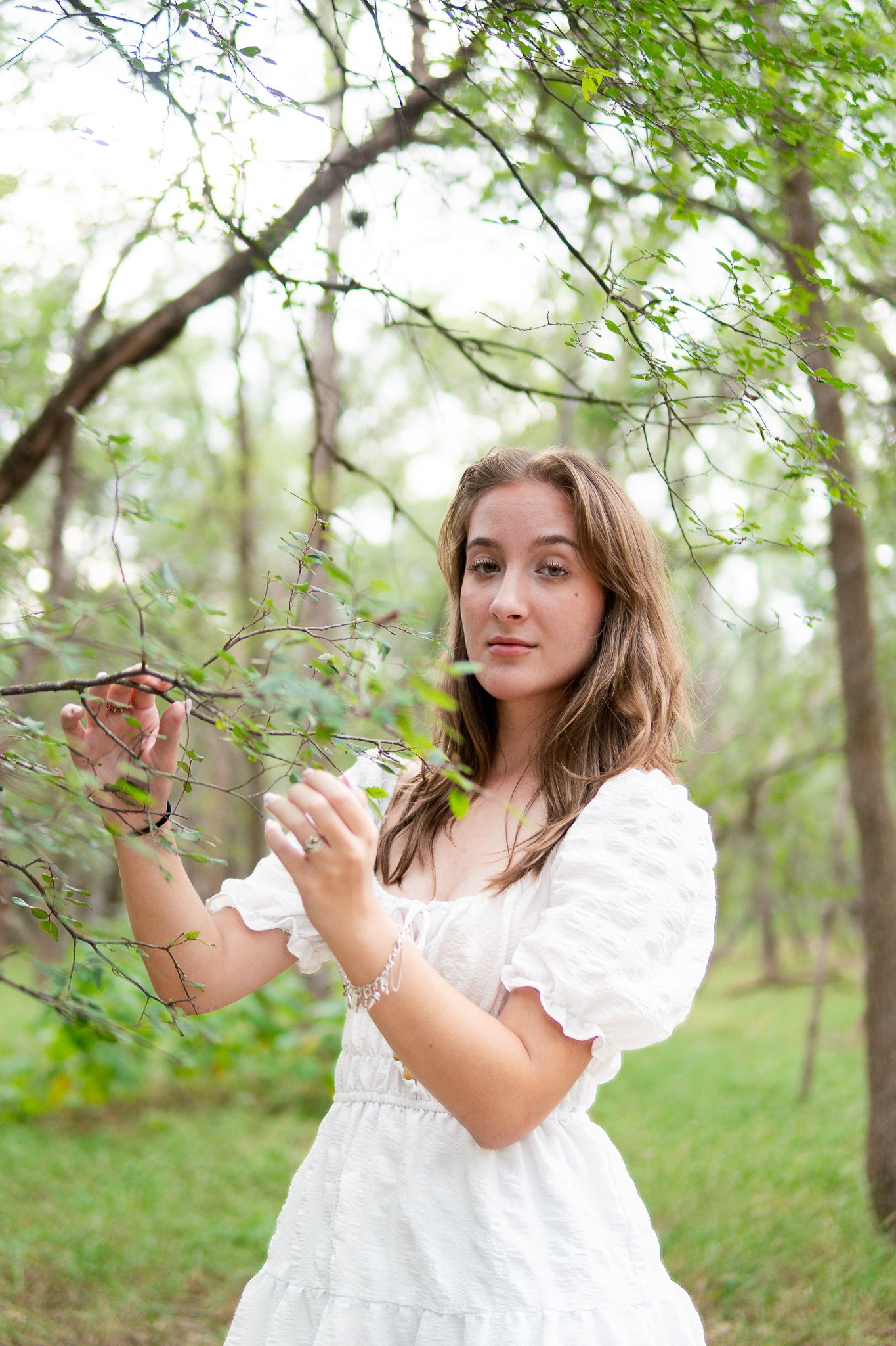 A person in a white dress stands in a forest setting among green leaves and branches on a spring day.