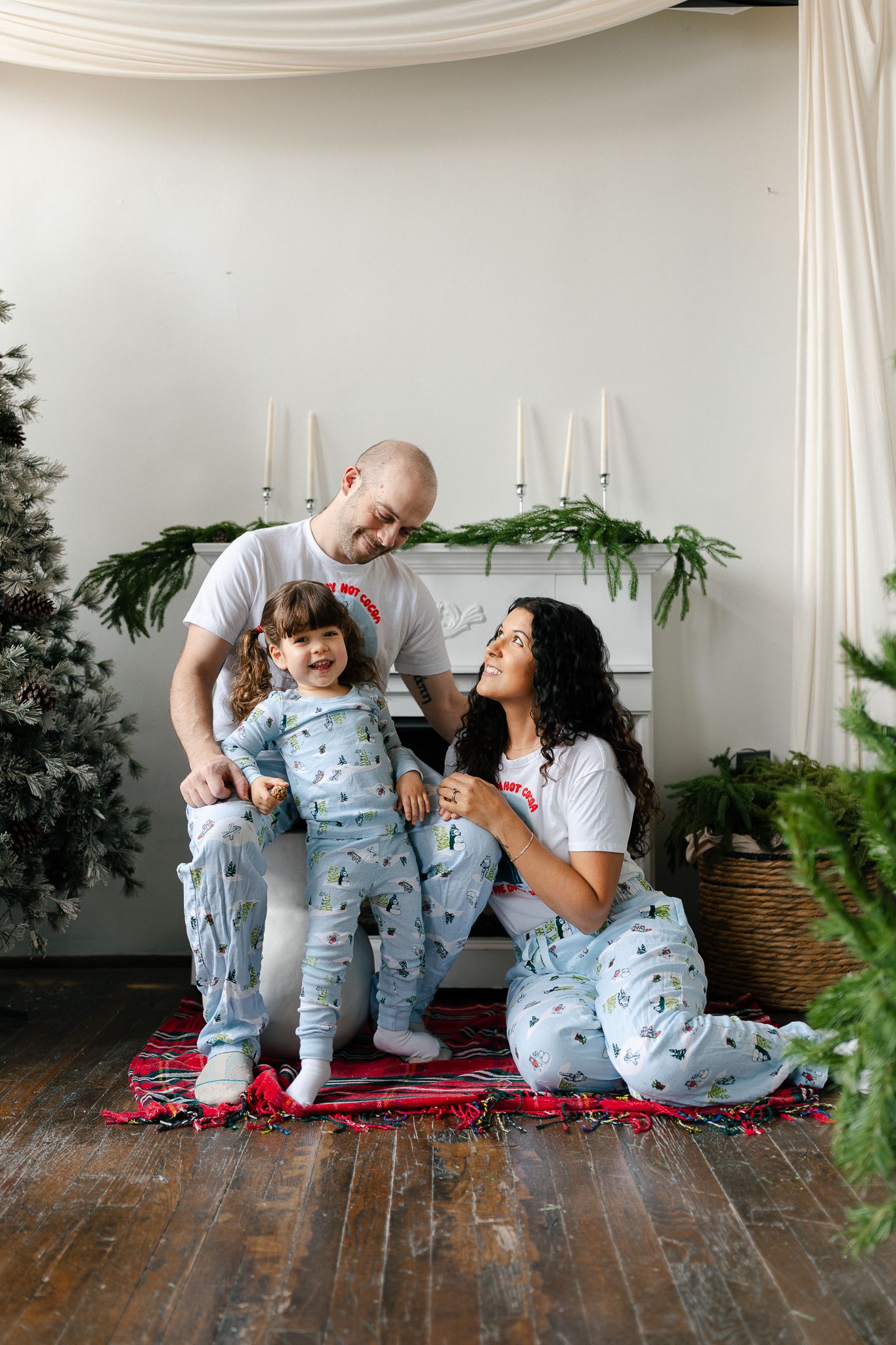Family wearing matching blue pajamas sits together on red blanket in bright white room with plants.