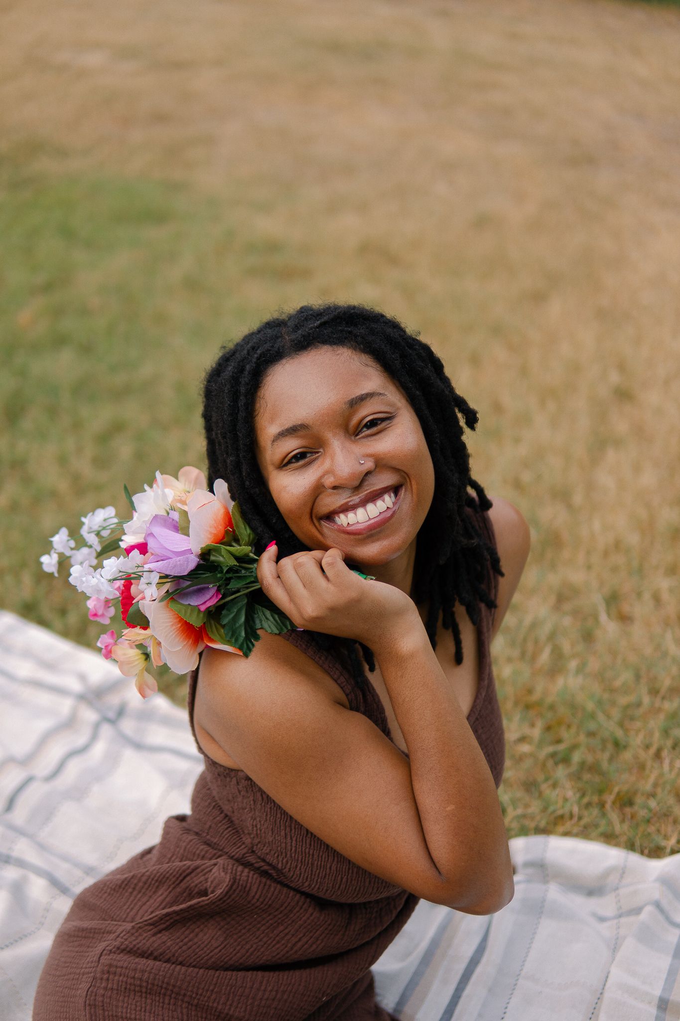 A person with dreadlocks smiles while lying on a striped blanket outdoors with pink flowers nearby.