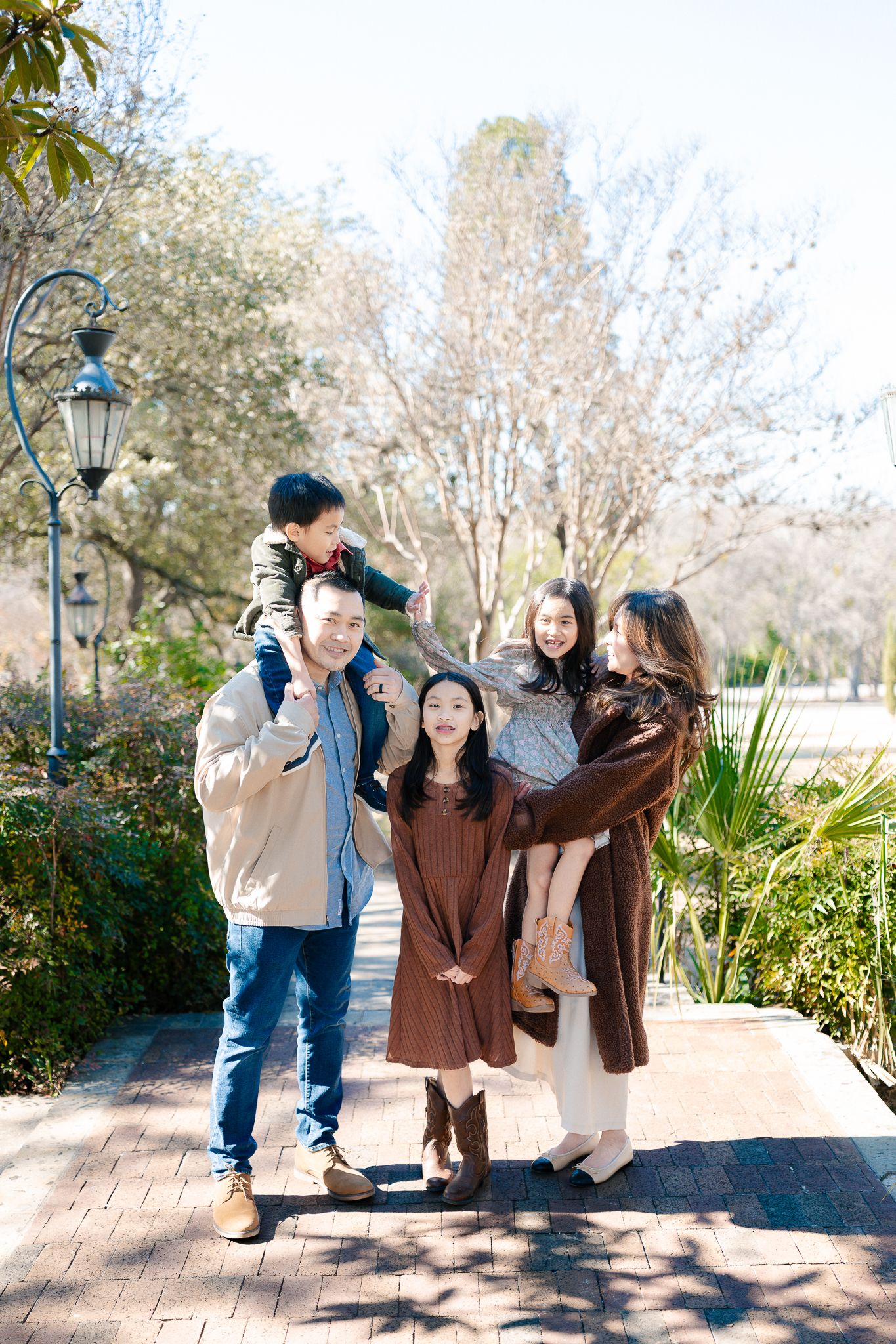 A group in coordinating brown outfits poses playfully together on a brick pathway surrounded by greenery.