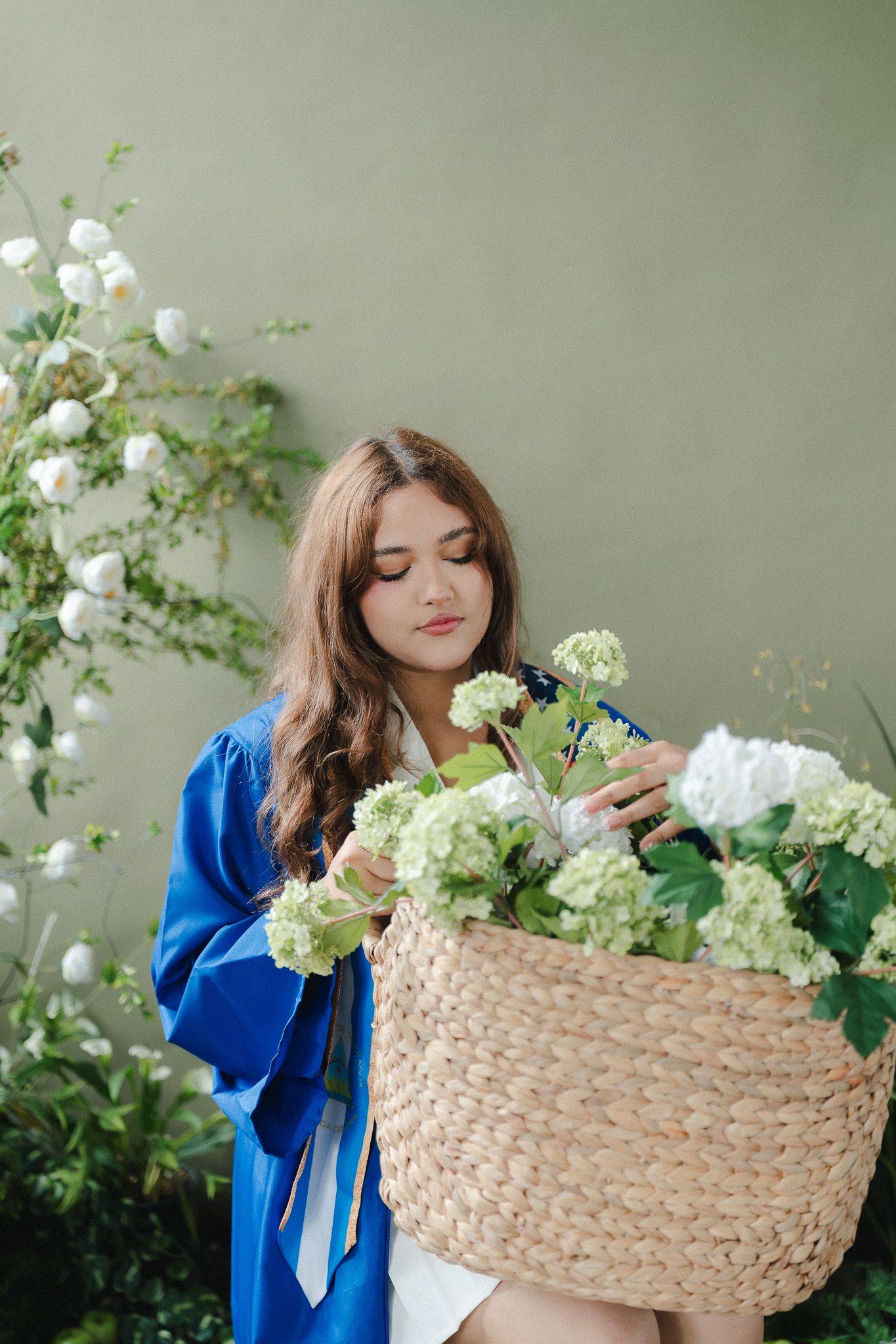 Young woman in blue graduation gown holding a wicker basket filled with white hydrangeas and flowers outdoors.