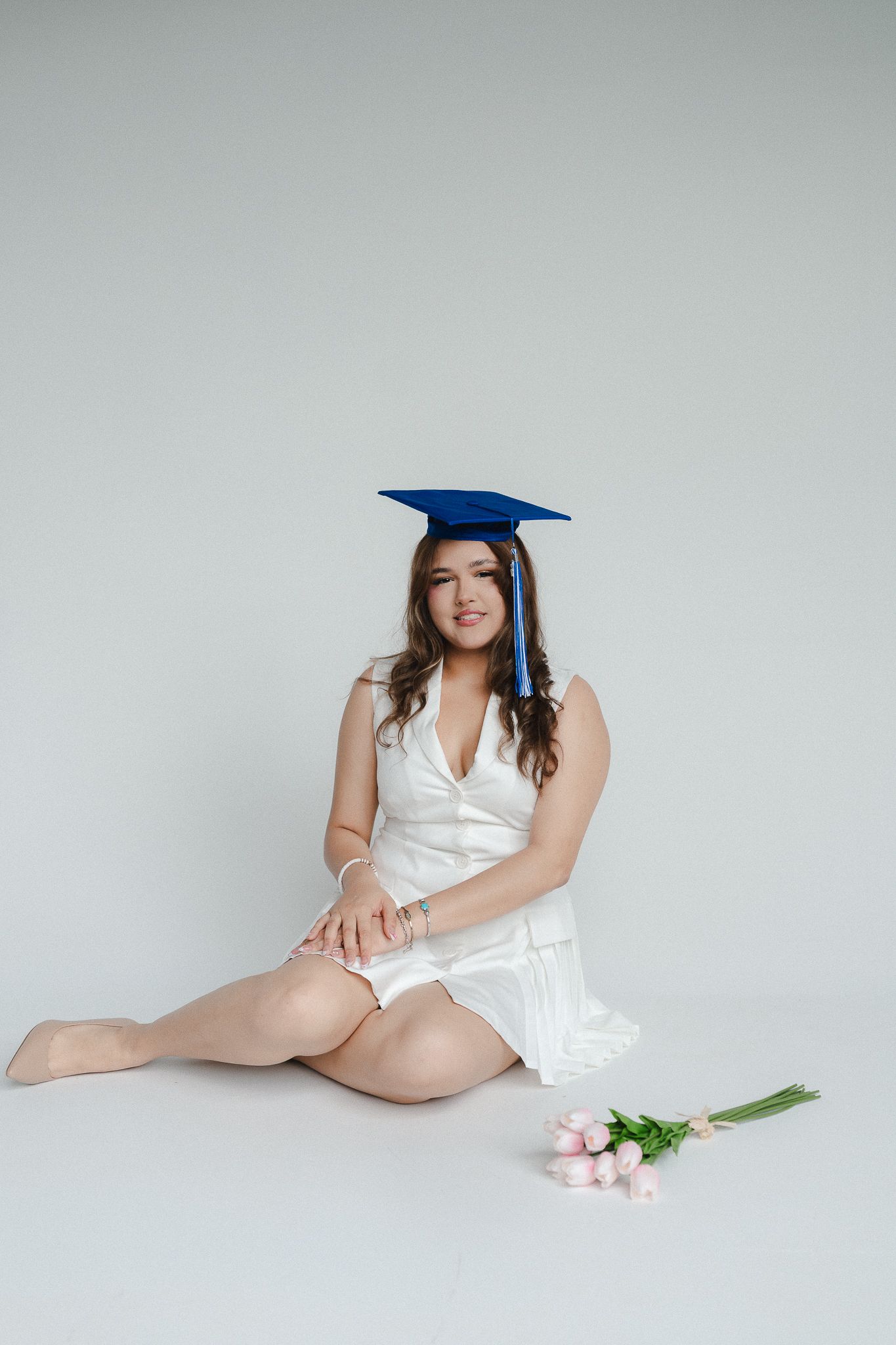 Young woman in white dress and blue graduation cap sits on floor with pink tulips, celebrating academic achievement.