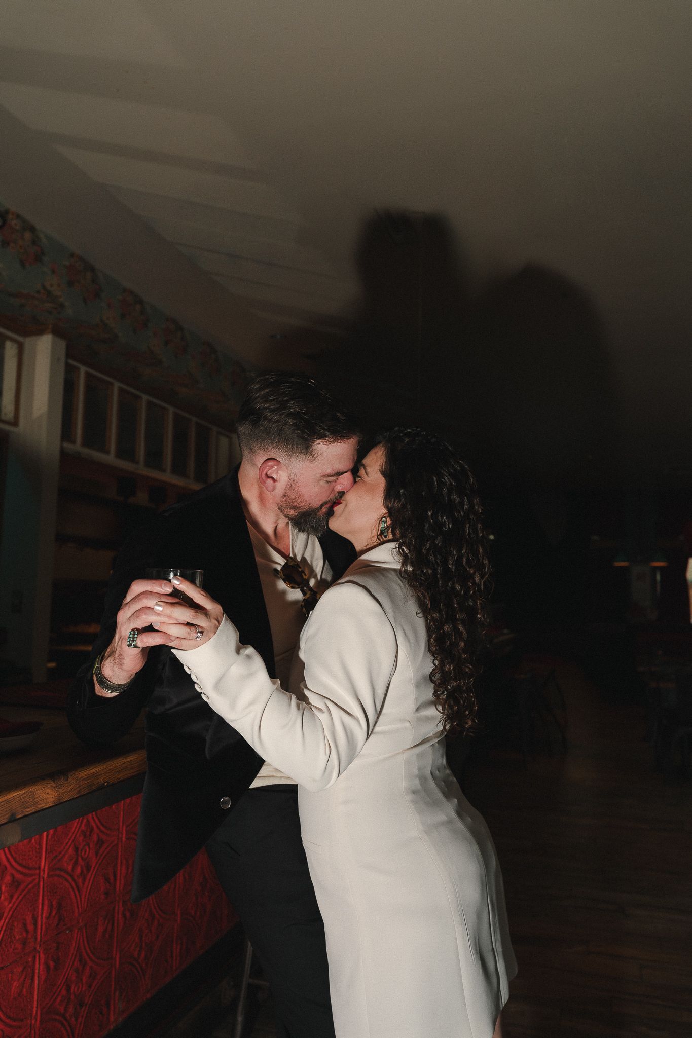 Couple sharing a romantic kiss while slow dancing in a dimly lit venue, woman in white blazer holding a drink.